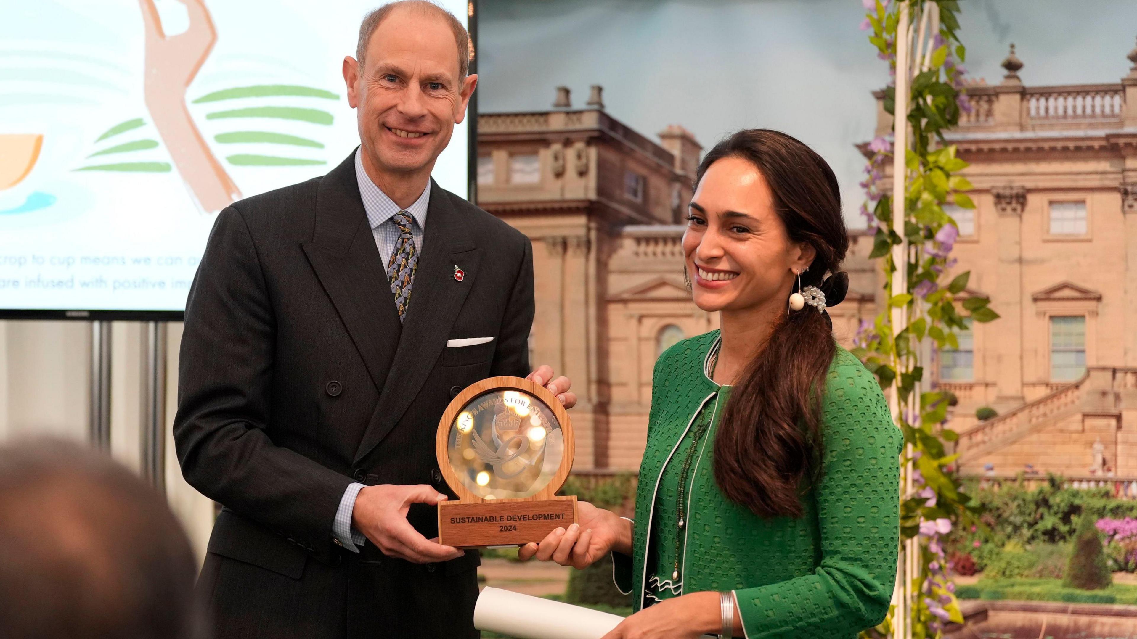 The Duke of Edinburgh, Prince Edward is smiling at the camera and wearing a black suit, blue shirt and patterned tie. Zahra Afshar is smiling at the camera, wearing a emerald green jacket and dress, with her dark hair pulled to one side. Both are holding a brown circular award.