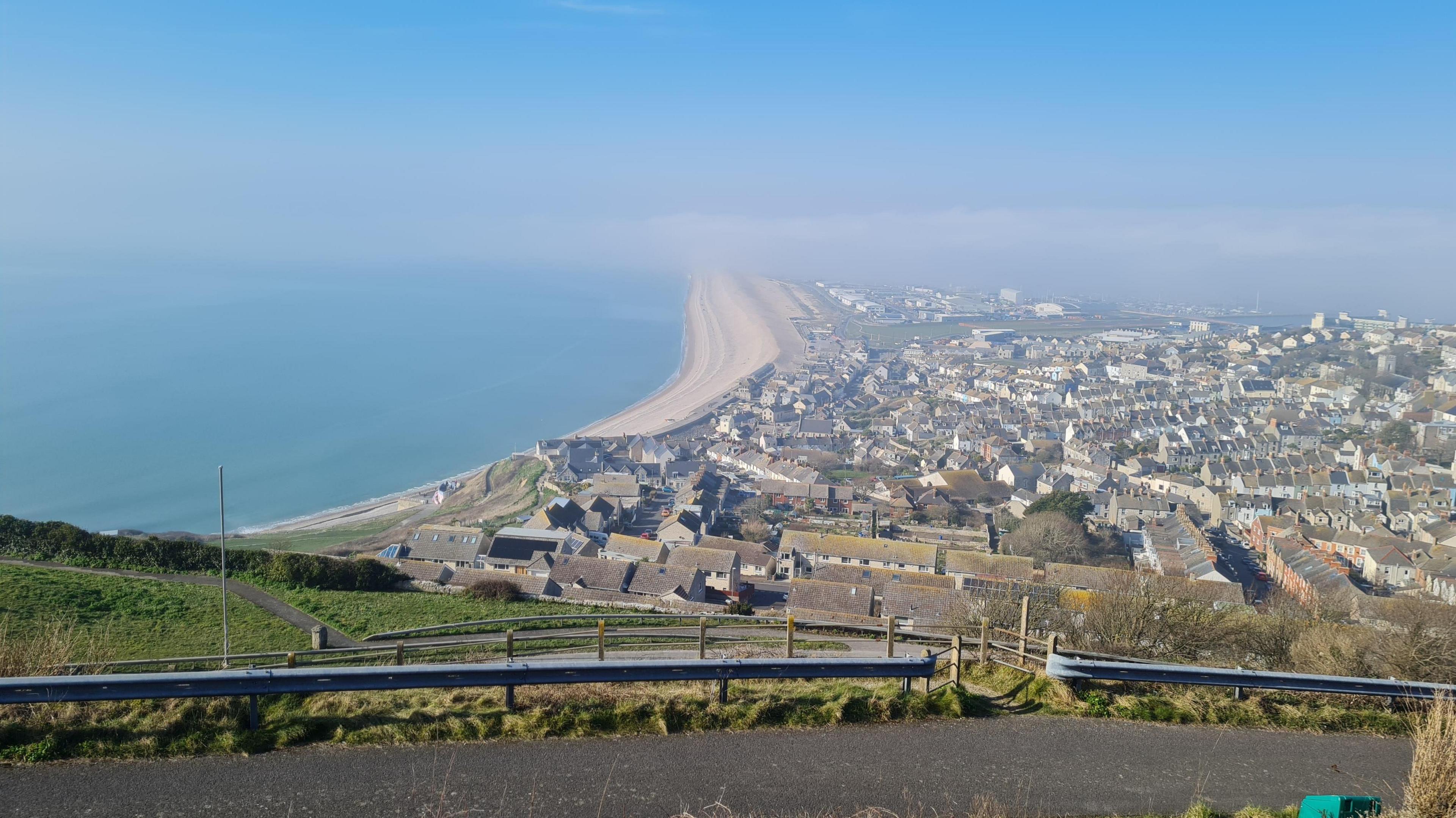 A photo taken from an elevated position on top of Portland looks out over the town and towards Weymouth that is shrouded in fog. The rest of the county has vanished into the clouds. In the foreground you can see homes and Chesil Beach where the sun is shining. 