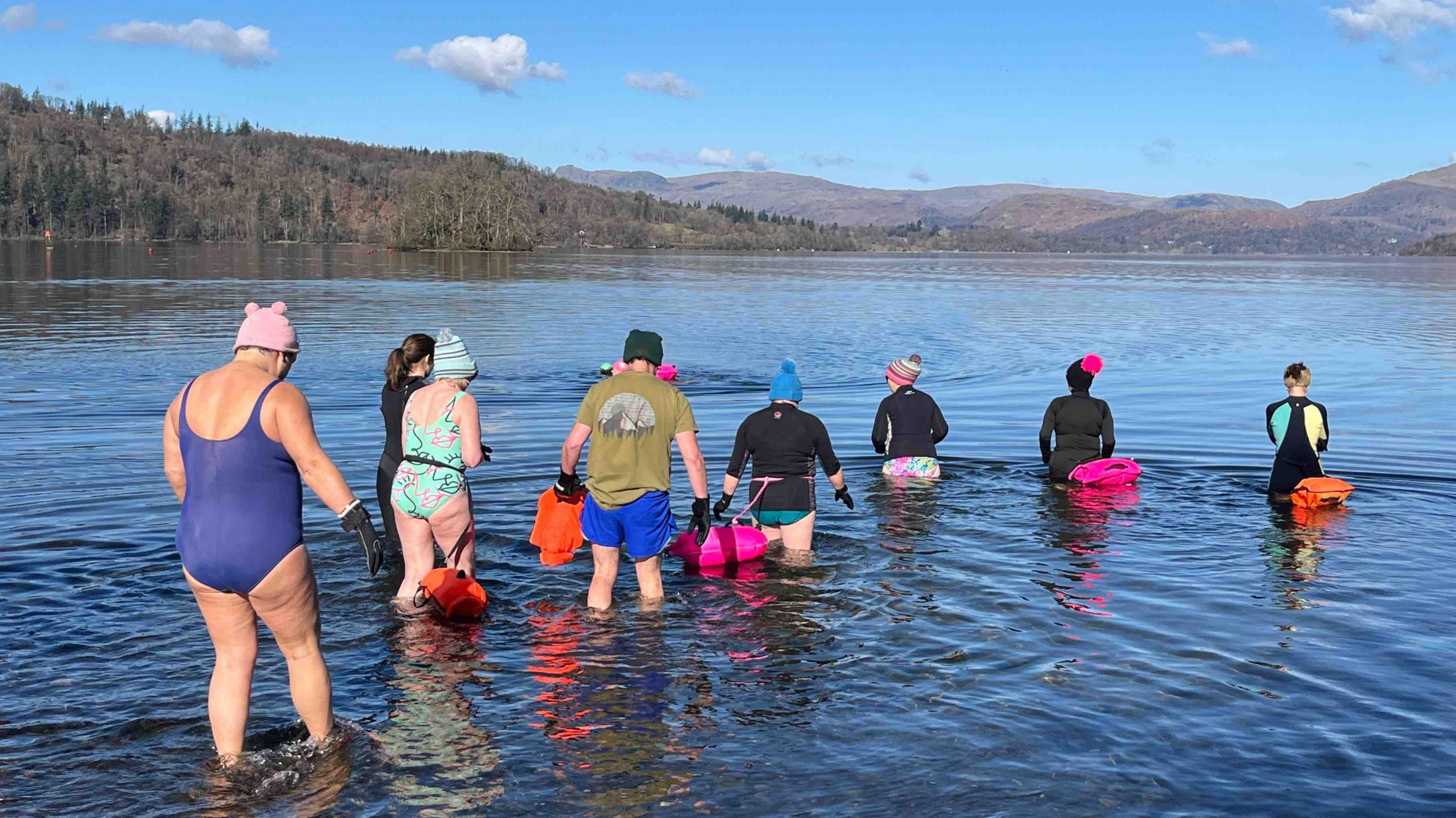 A group of swimmers with their backs turned enter the lake wearing colourful costumes and bathing hats - they make patterns in the dark water as they enter. In the distance you can see bare brown coloured mountains and conifer trees on the lake's banks.