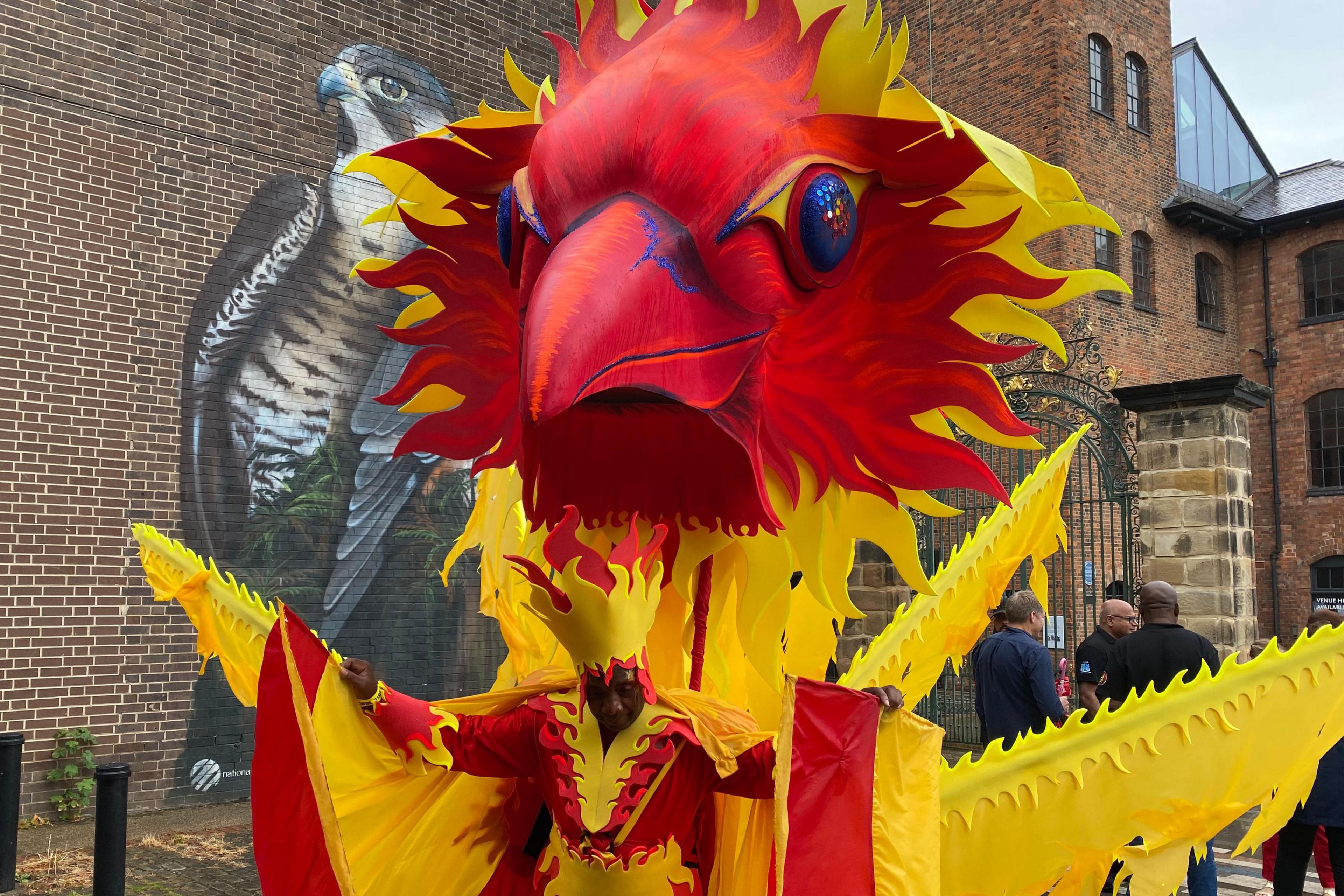 A man in a huge phoenix outfit next to the Silk Mill and its peregrine falcon mural