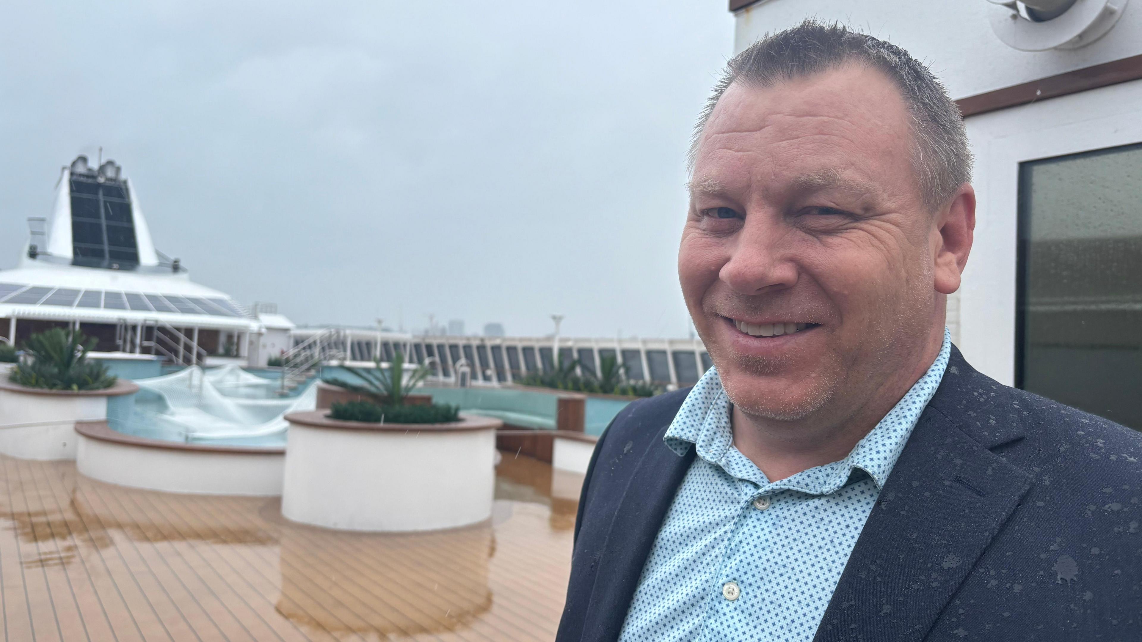 Mikael Petterson stands on a wet cruise ship deck. He has short grey hair, a blue shirt and jacket and is covered in rain. 