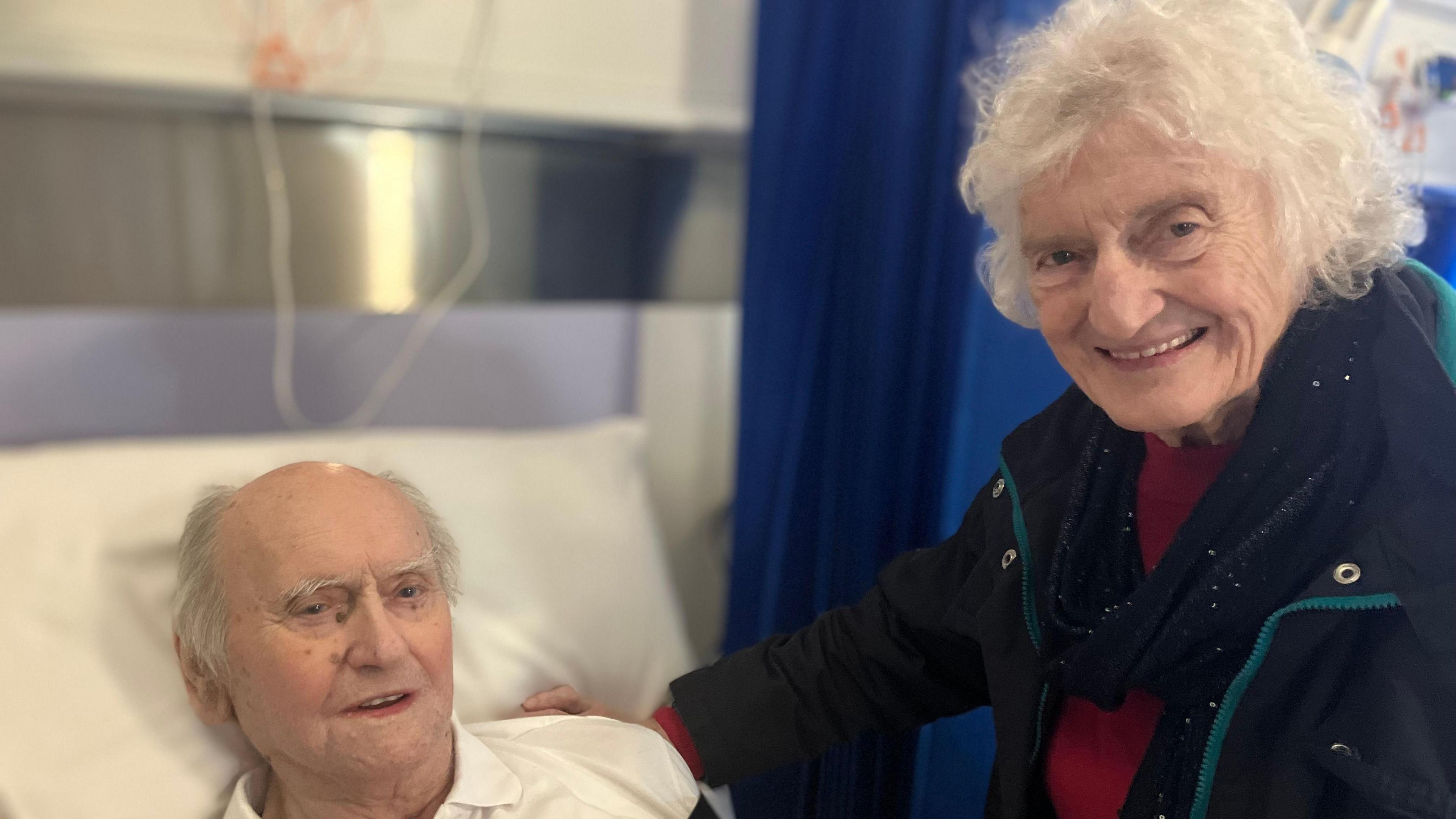 Clive Donavan is elderly, has grey hair and is in a hospital bed inside the pod. His wife Anne has grey hair and is wearing a navy coat and red jumper. 