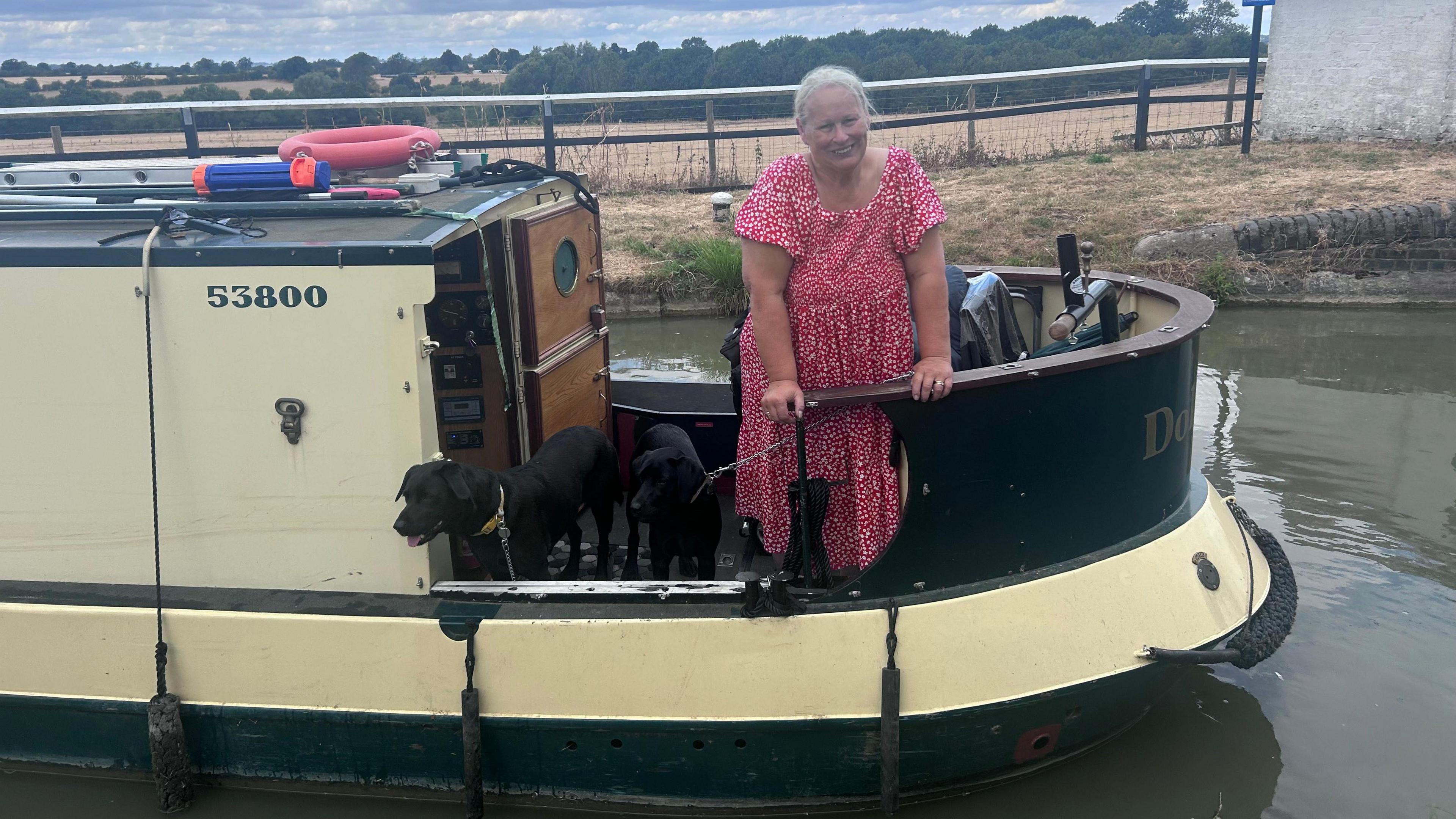 A woman standing on a canal boat. She is smiling and has her long hair tied back. She is wearing a red dress. The boat is on water with fields behind her. 