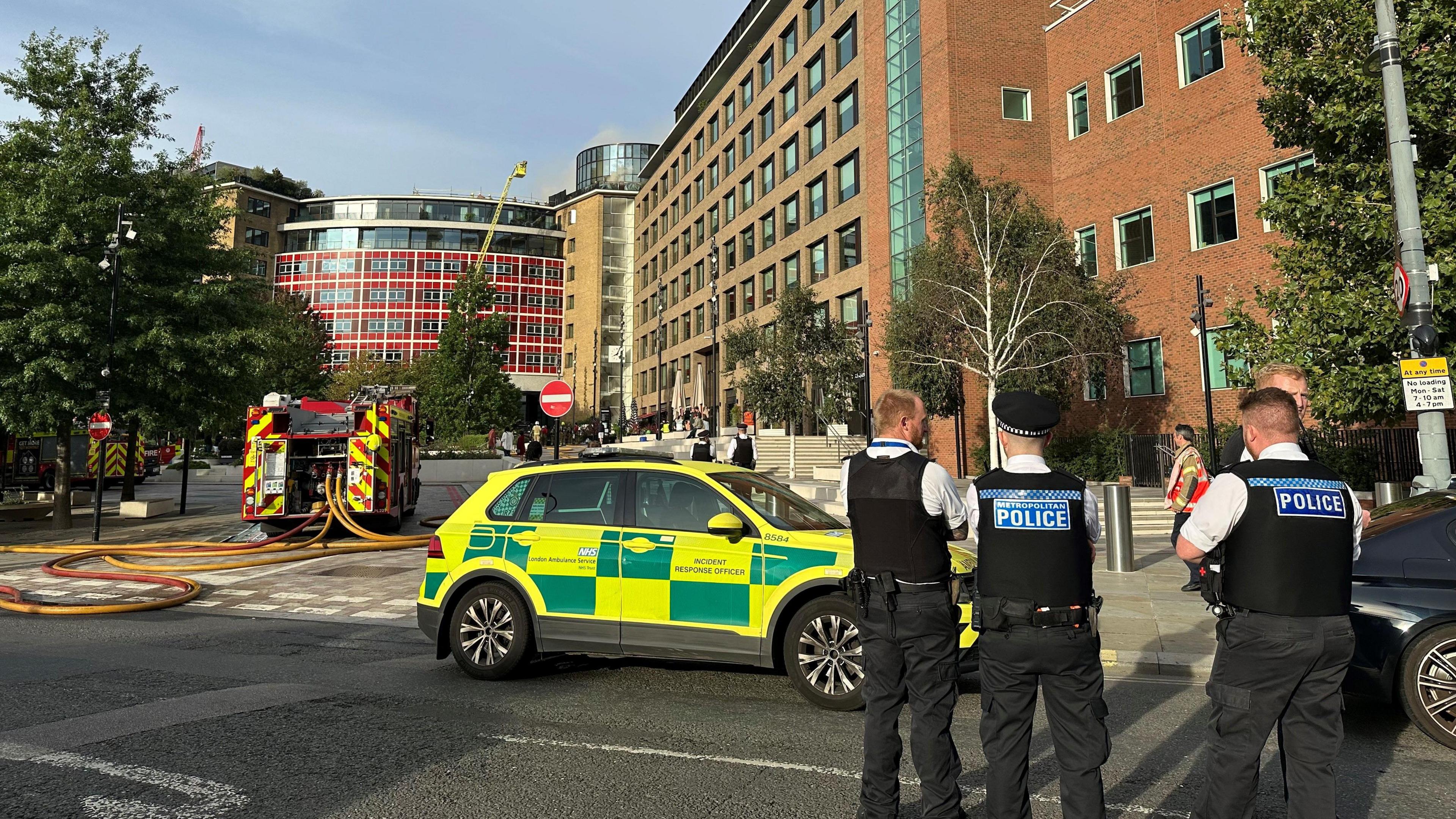 Police officers stand in the road facing the building. A London Ambulance car is parked on the road, and a fire engine is seen in the background. 