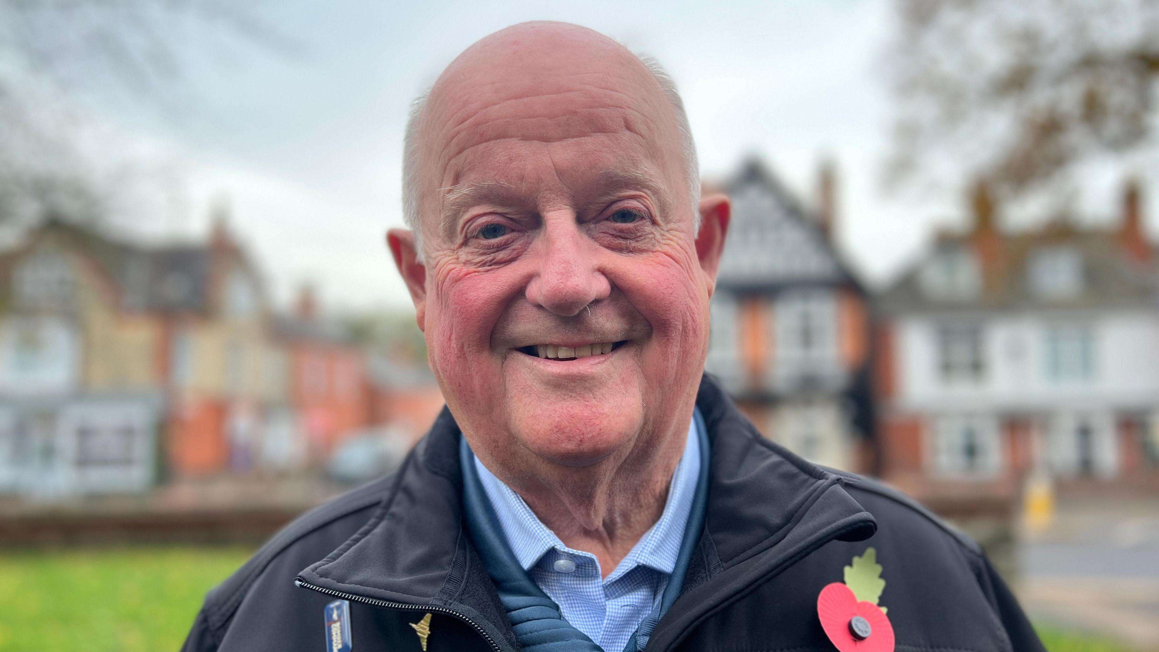 Man with blue shirt, blue jumper and black coat wearing a red poppy badge. He is smiling. There are houses behind him.