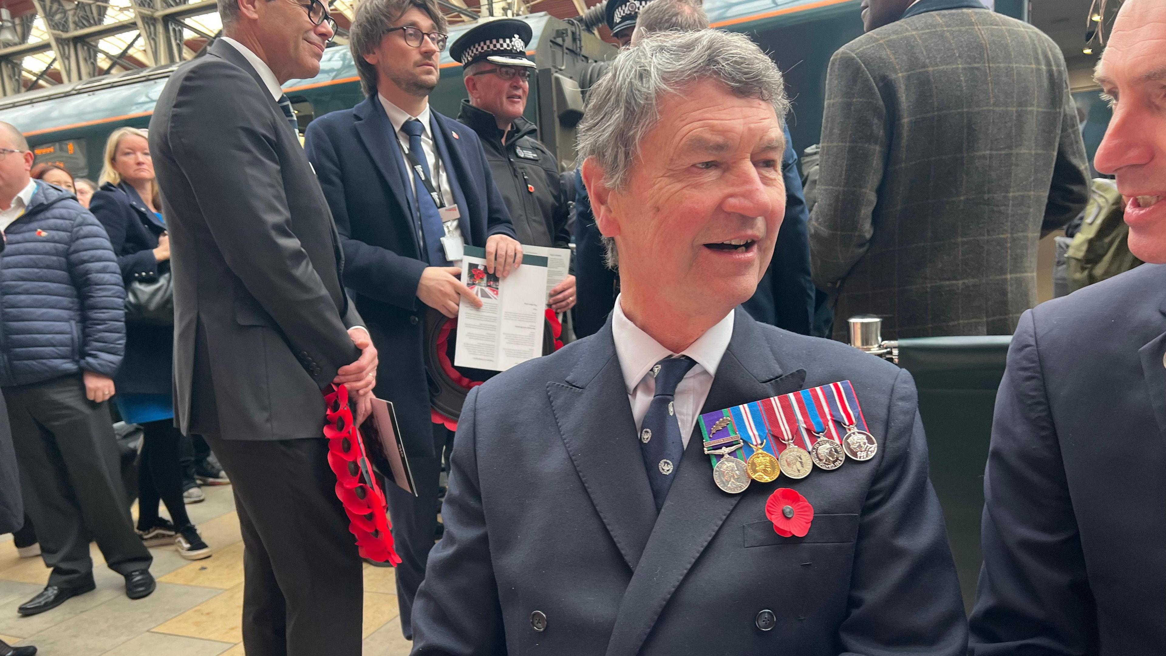 Vice Admiral Sir Tim Lawrence sits on a bench on a railway platform with people in the background. He wears a navy blue suit and tie with a white shirt. He has medals on the chest of his jacket and a poppy underneath.