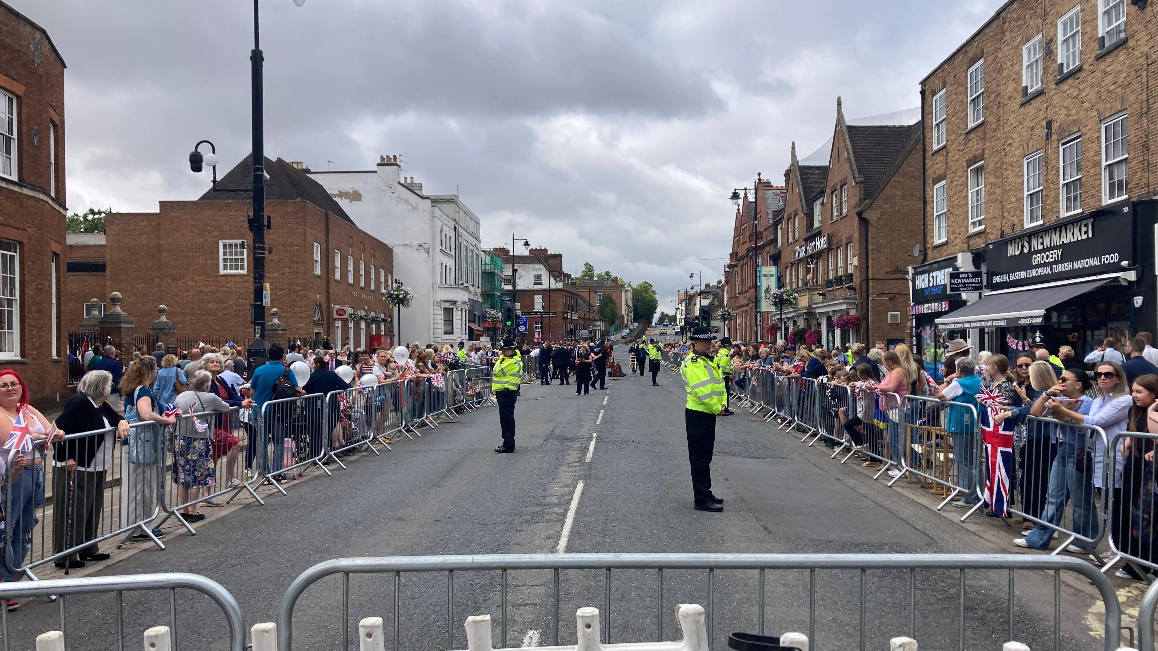 Crowds of being line Newmarket High Street and stand behind barriers. Uniformed officers stand in the middle of the road watching the crowds