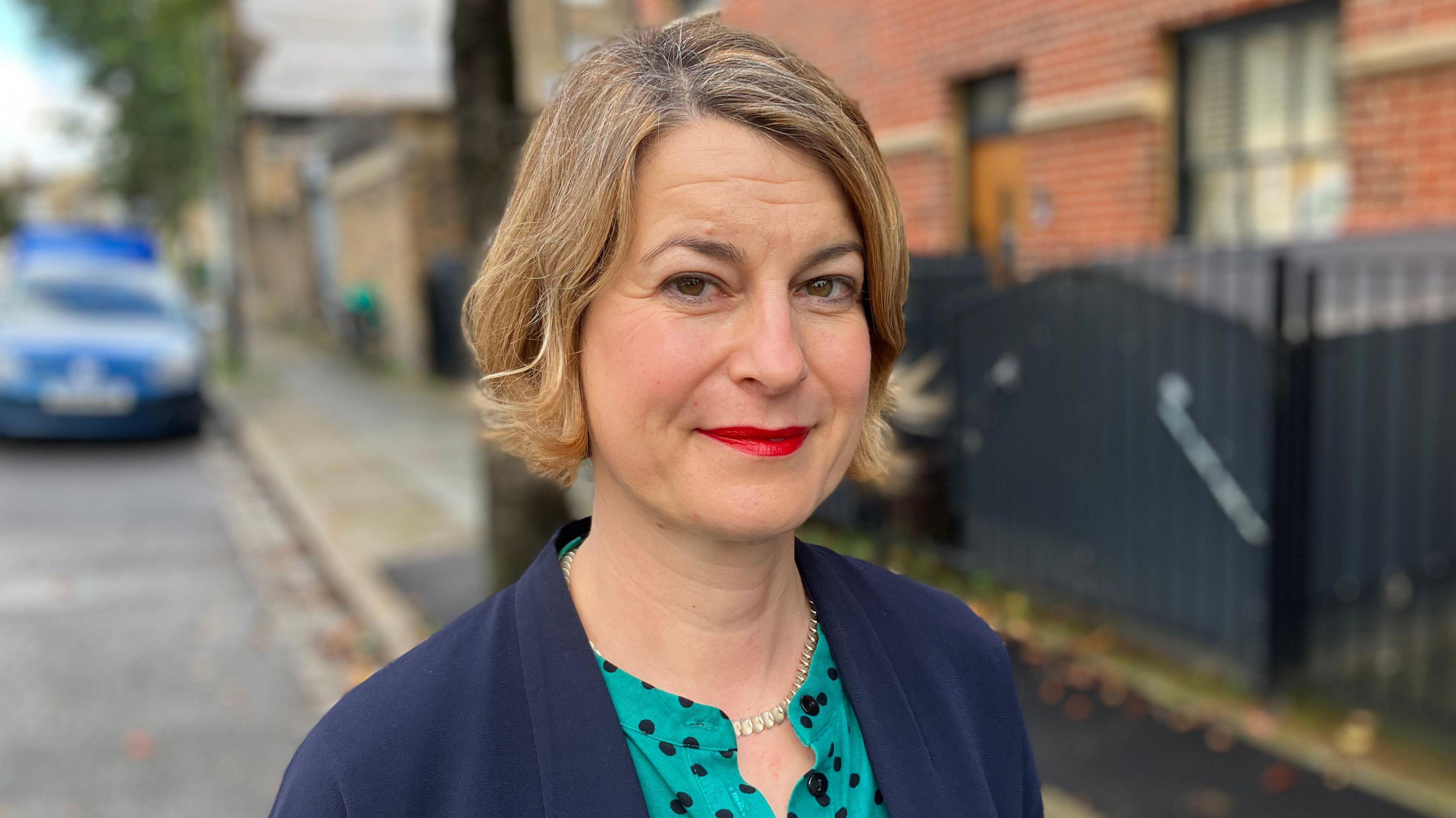 Helen Hayes, the MP for Dulwich and West Norwood. She is standing outside on a residential street which has housing and cars parked on the side of the road. She is looking directly at the camera and is smiling. She is wearing a teal and black polka dot blouse with a navy tailored jacket over the top. 