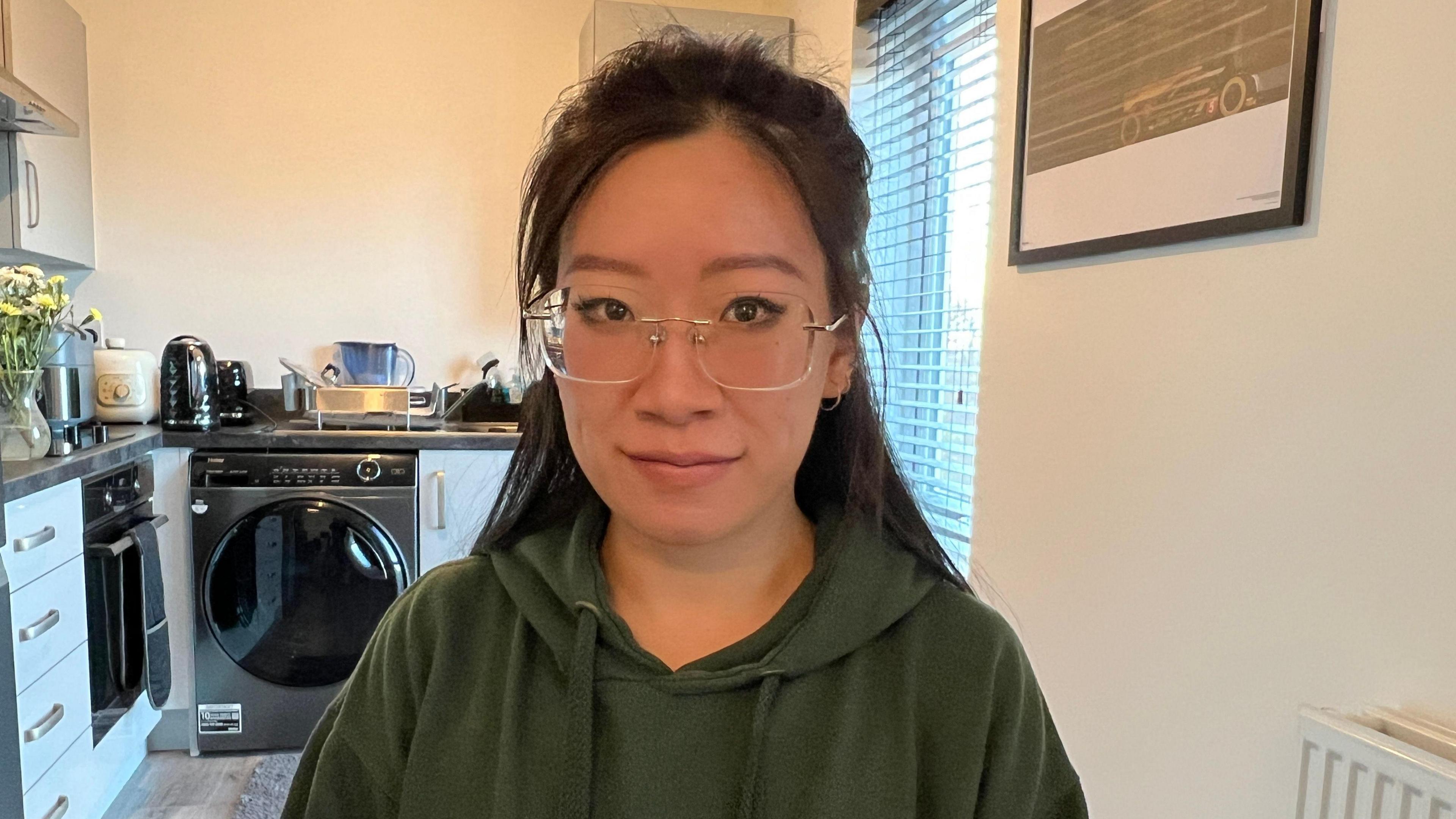 A woman in a kitchen looks at the camera with a neutral expression. She wears frameless glasses and a green hoodie and her dark hair hangs behind her shoulders. Behind her is a washing machine to the left and a vase of yellow flowers is on the worktop beside it.

