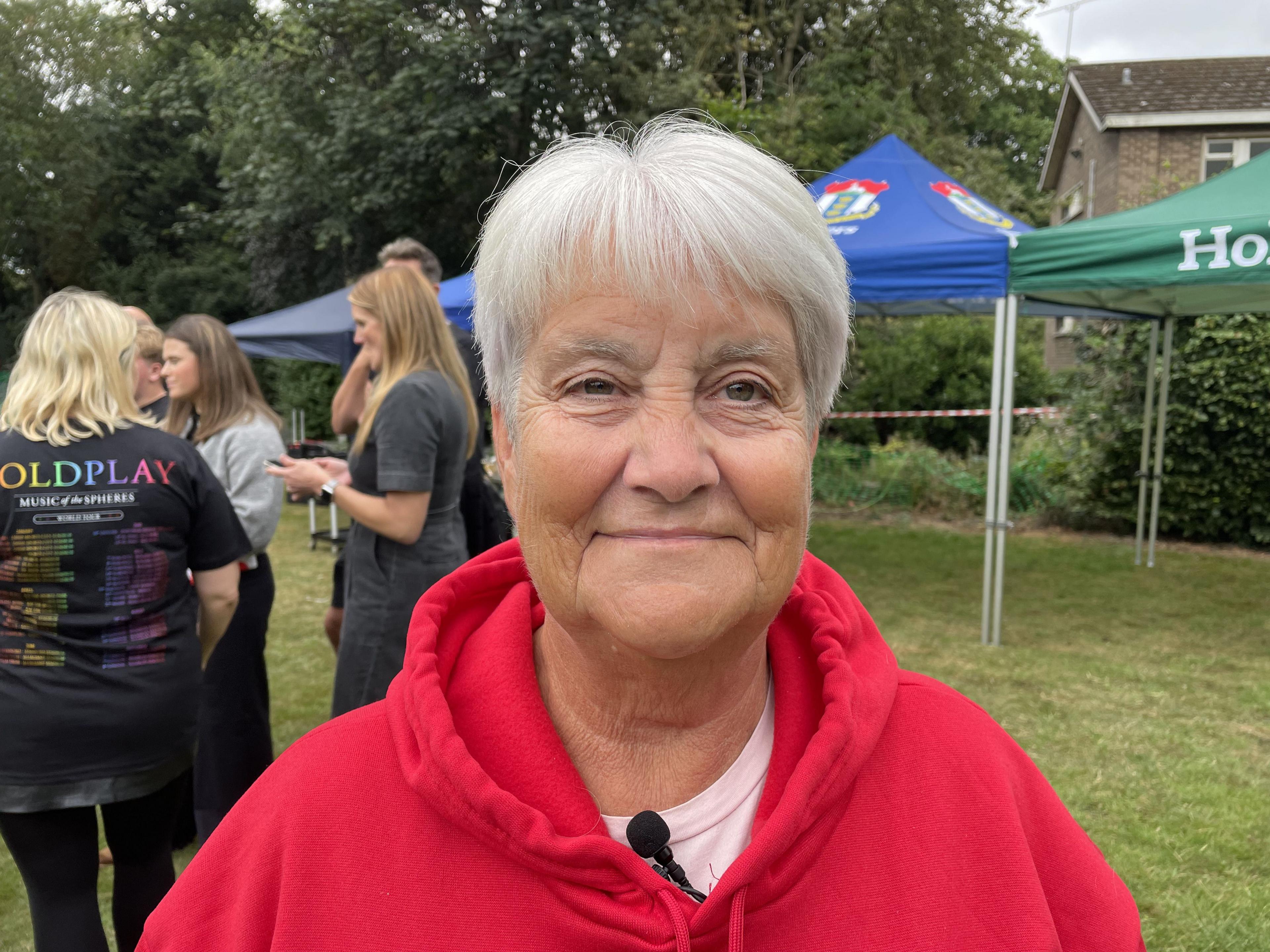 Woman with short grey hair, red hoody and pink top. She is stood on grass and there is a bush and a marquee behind her as well as people.