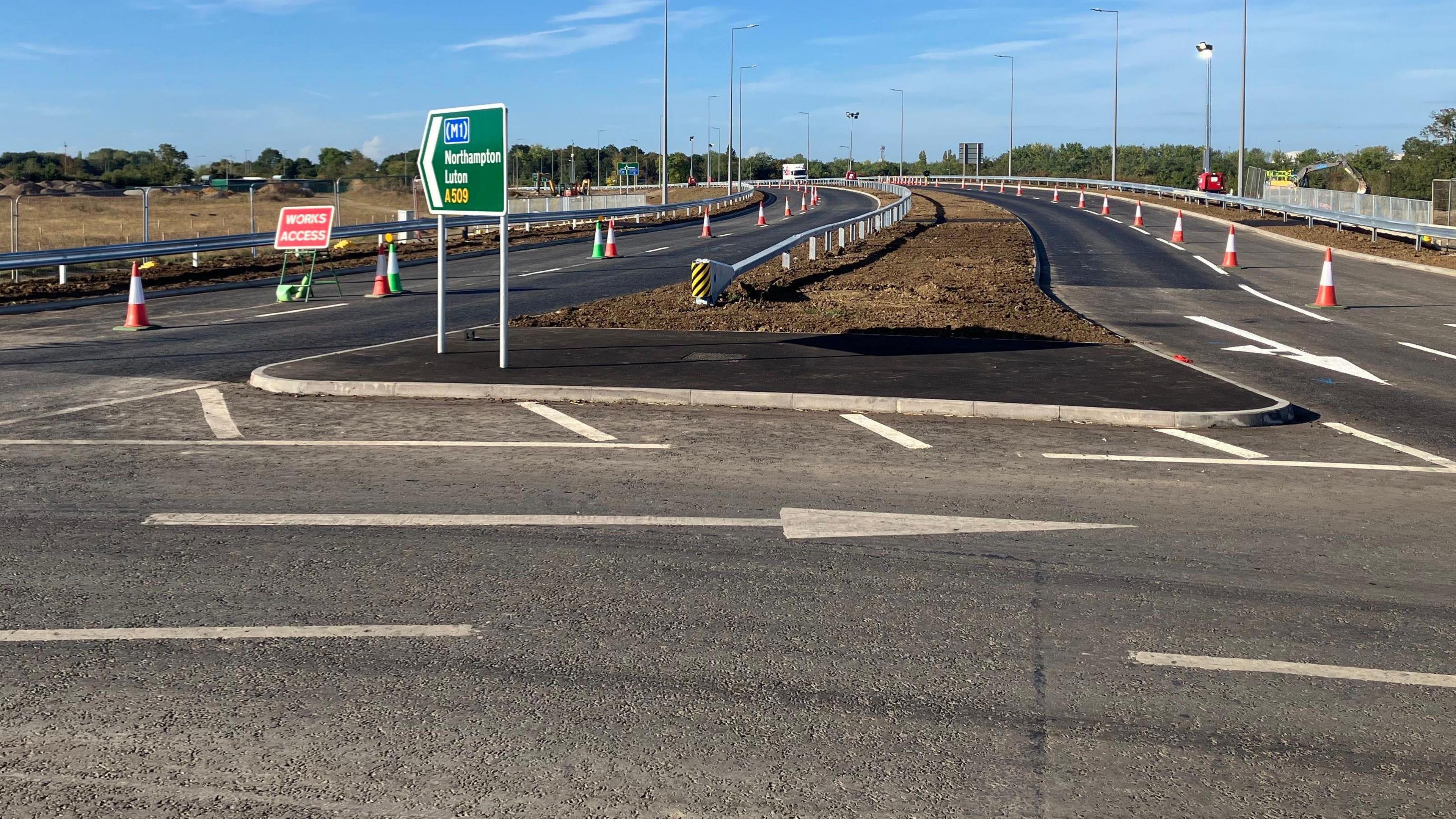 A picture of the newly reopened A509. You can see the road which still has some traffic cones on it, plus a sign that says M1, Northampton and Luton on it.