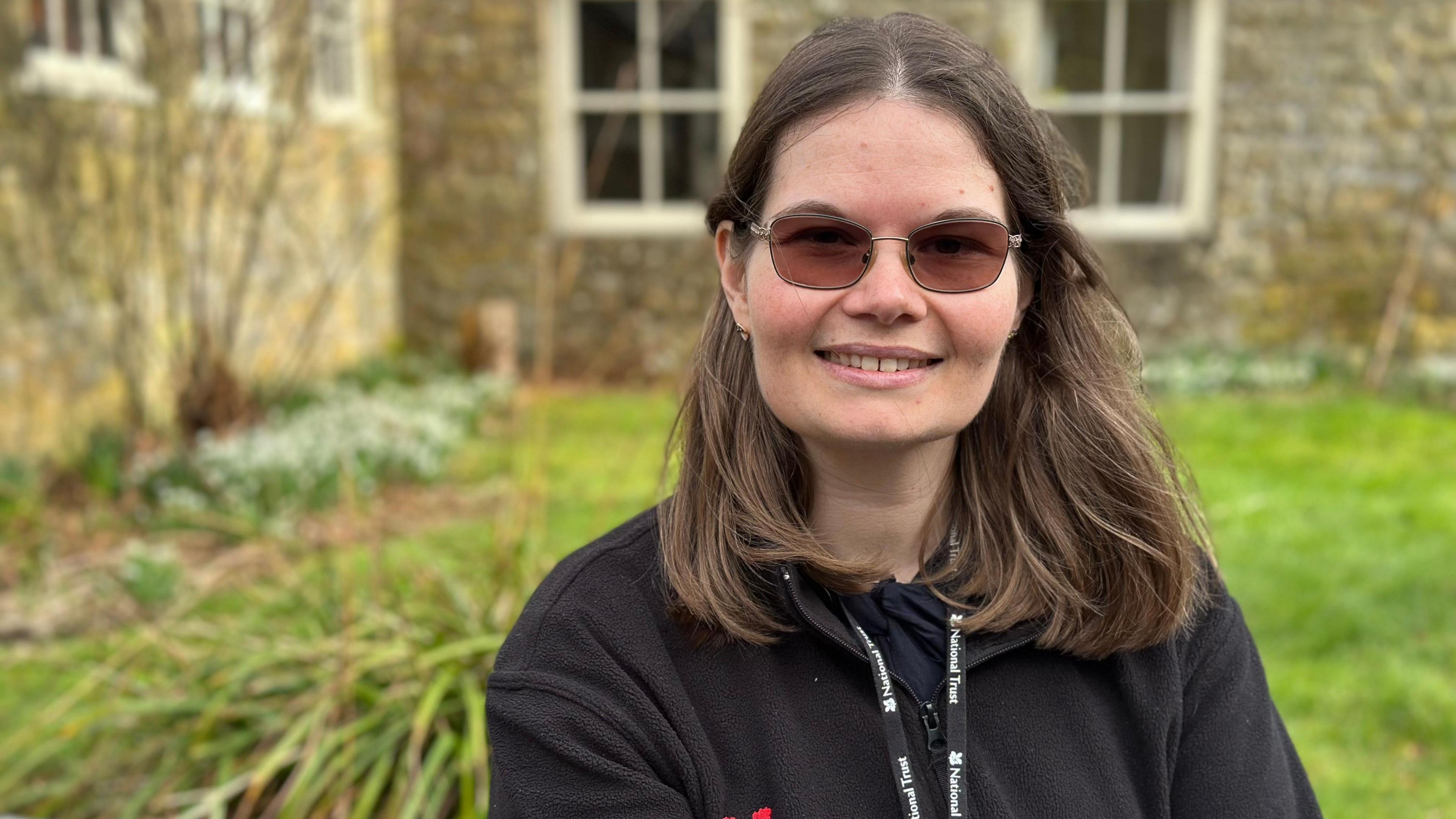 Natalie Warren is smiling at the camera. She has medium-length straight brown hair and is wearing glasses, a black fleece and a lanyard. 