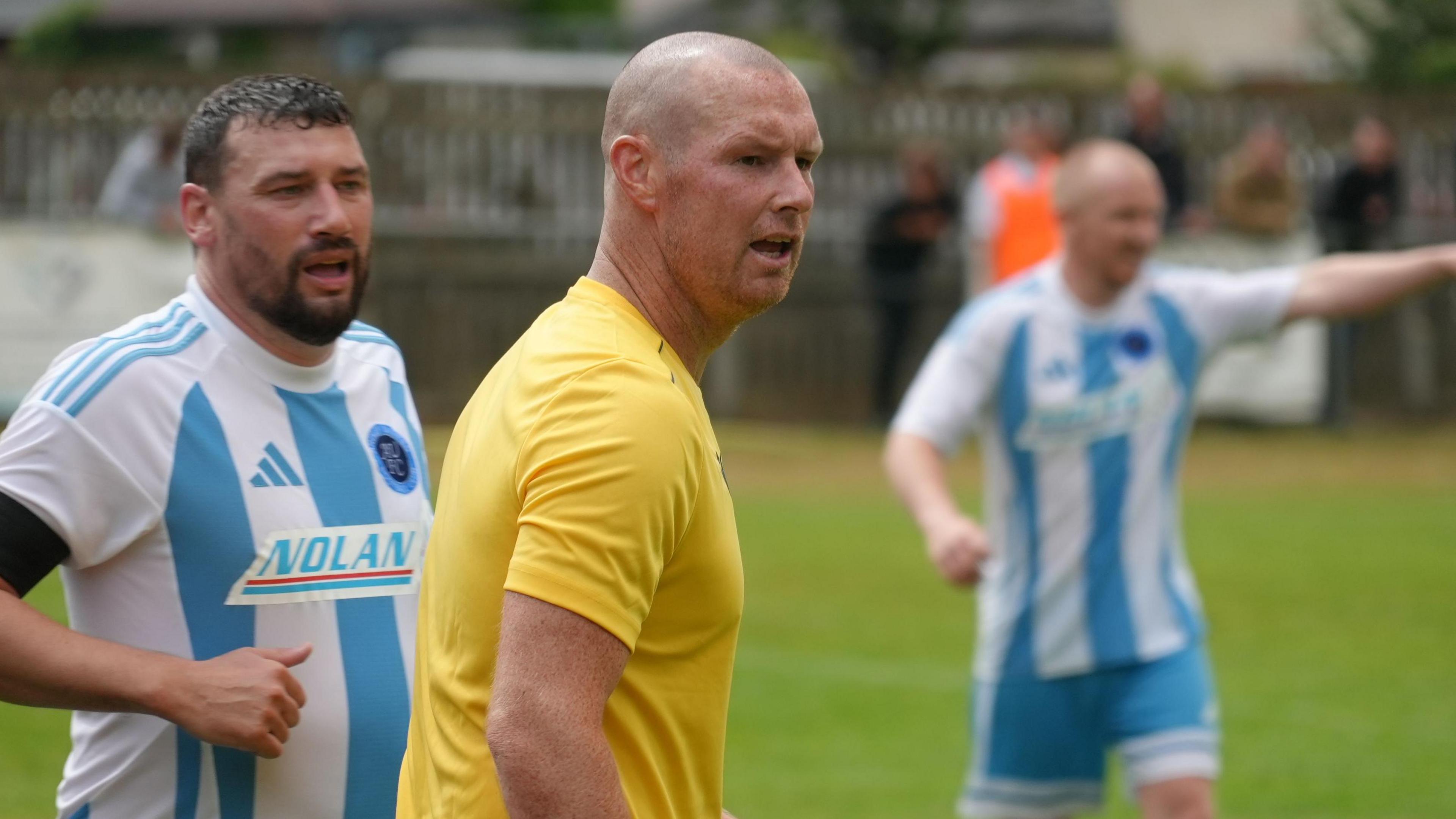 James Constable has a bald head and is wearing a yellow Oxford United strip. He is being marked by an Ardley player wearing a blue and white striped shirt.