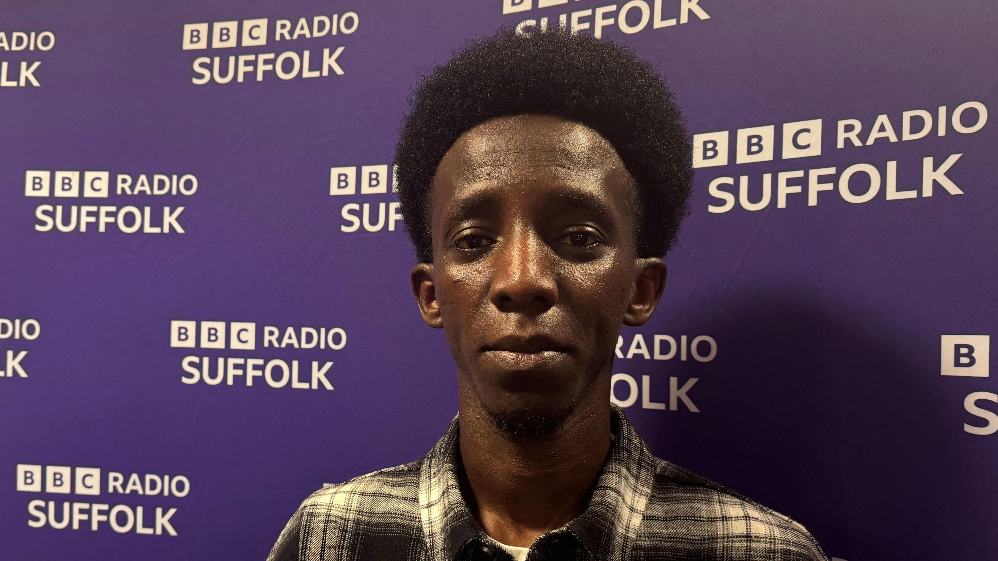 A young man wearing a black and white shirt while standing in front of a purple BBC Radio Suffolk backdrop. He is looking into the camera.