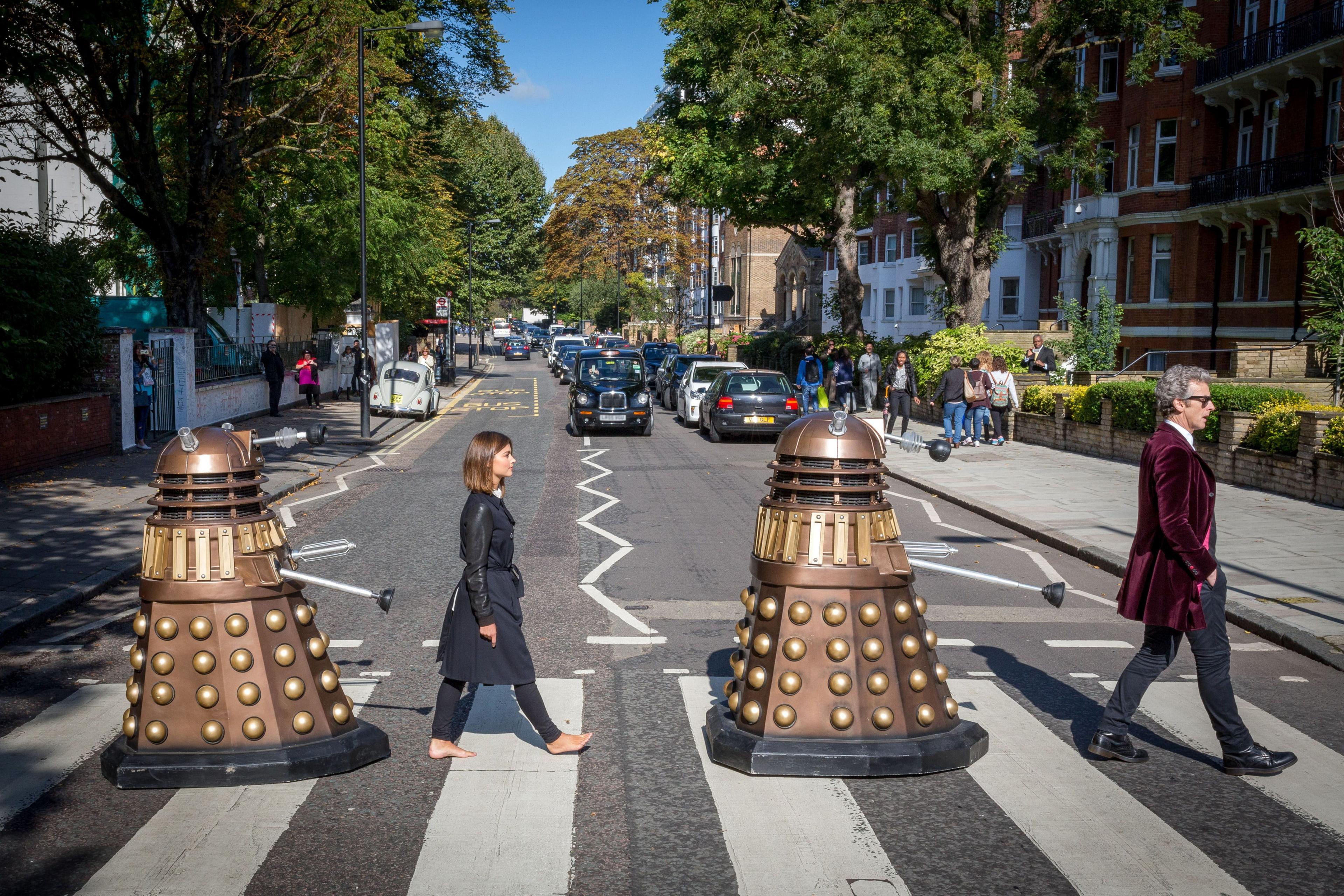 Publicity image for series 9 with Jenna Coleman (barefoot) and Peter Capaldi with 2 daleks on Abbey Road zebra crossing.