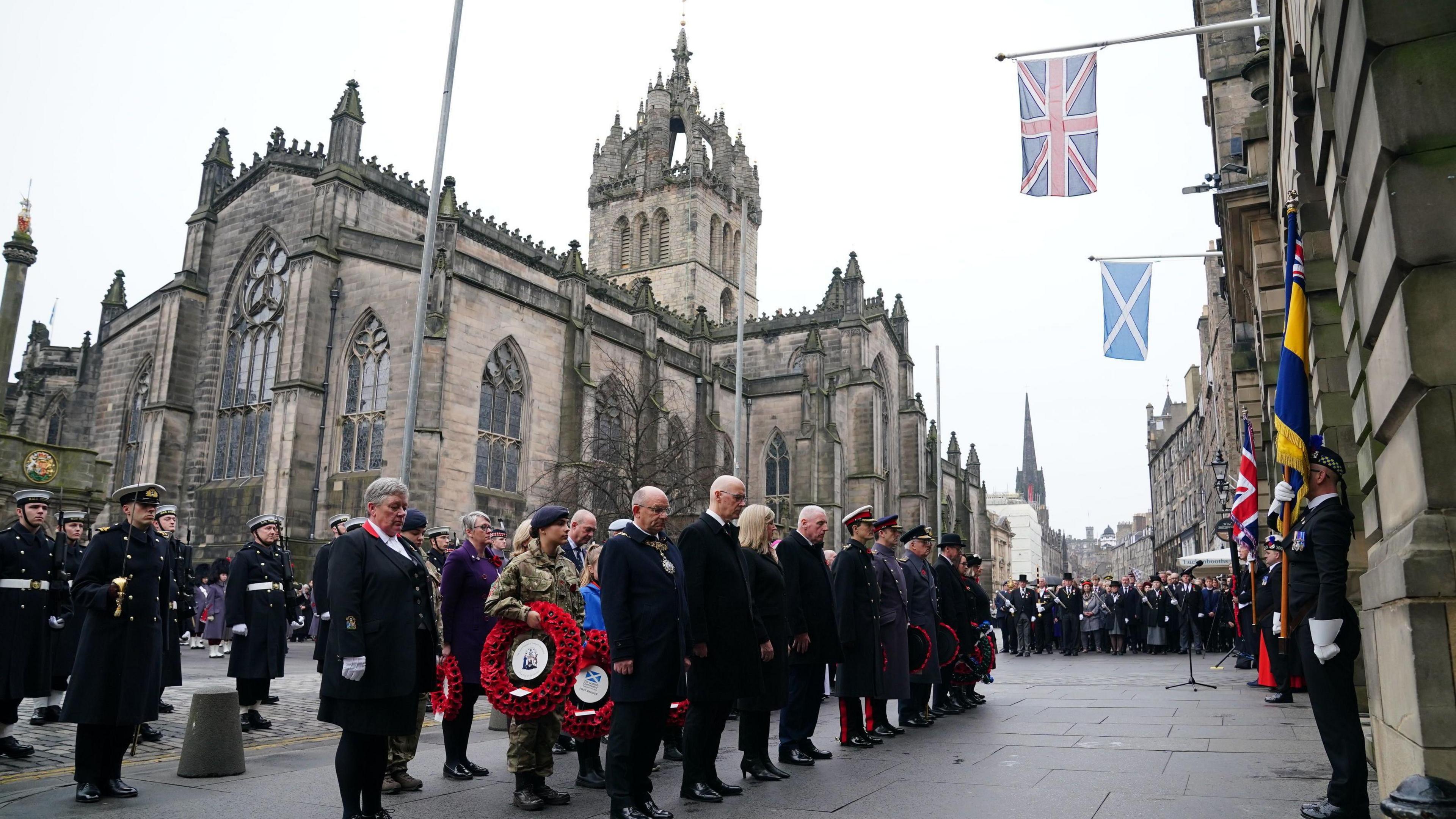 First Minister John Swinney with the Lord Provost of Edinburgh and service personnel during the two minutes' silence outside the city chambers where flags are flying. Some are holding poppy wreaths. St Giles Cathedral is behind them and there are people further up the Royal Mile.