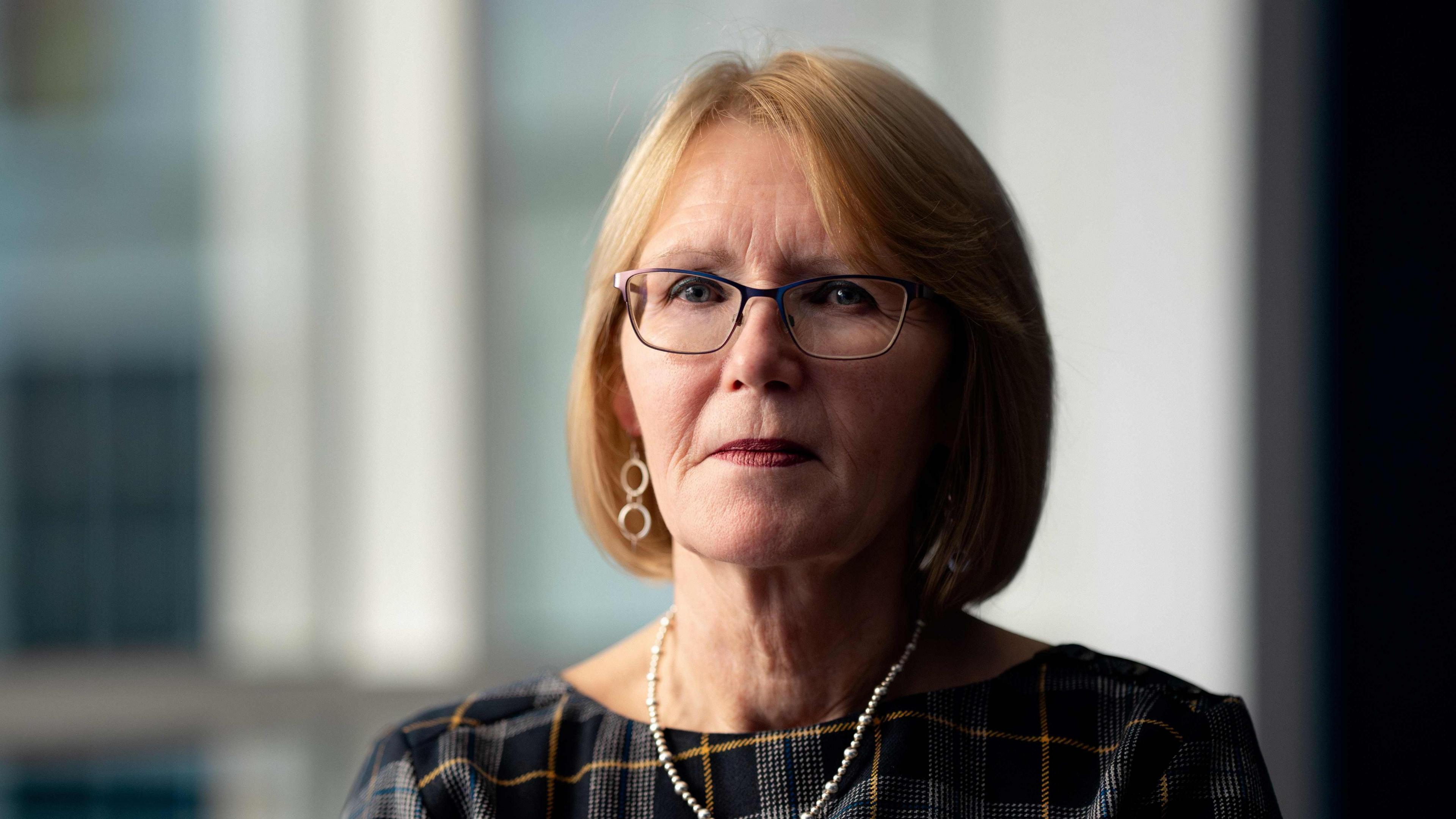 Image of Teresa Medhurst, chief executive of the SPS, a middle-aged woman with pale red hair in a bob, small brown-rimmed glasses and silver earrings and necklace. She wears a smart checked dress in black, white and yellow and is sitting in an office of some kind, a blurred view of large windows behind her.