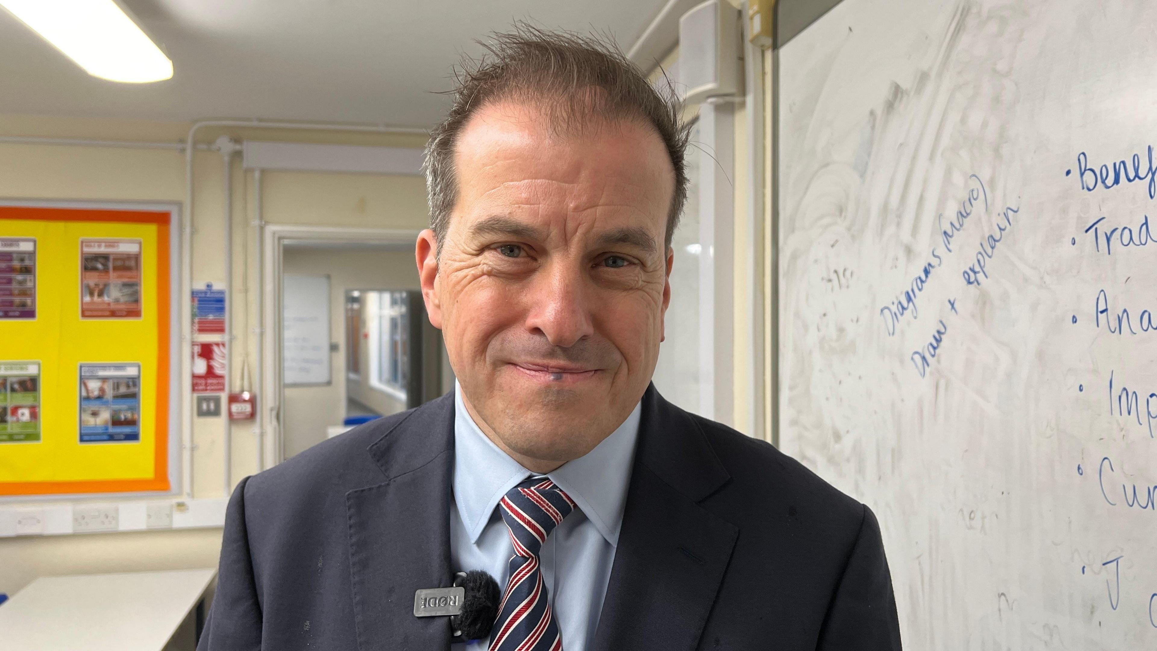 Rob Pearsall wearing a light blue collared shirt, a red, blue and white striped tie and a dark blazer. He is standing in a classroom beside a dirty whiteboard and is smiling at the camera, with a mic clipped to his lapel. Behind him there is a bright yellow wall display. 