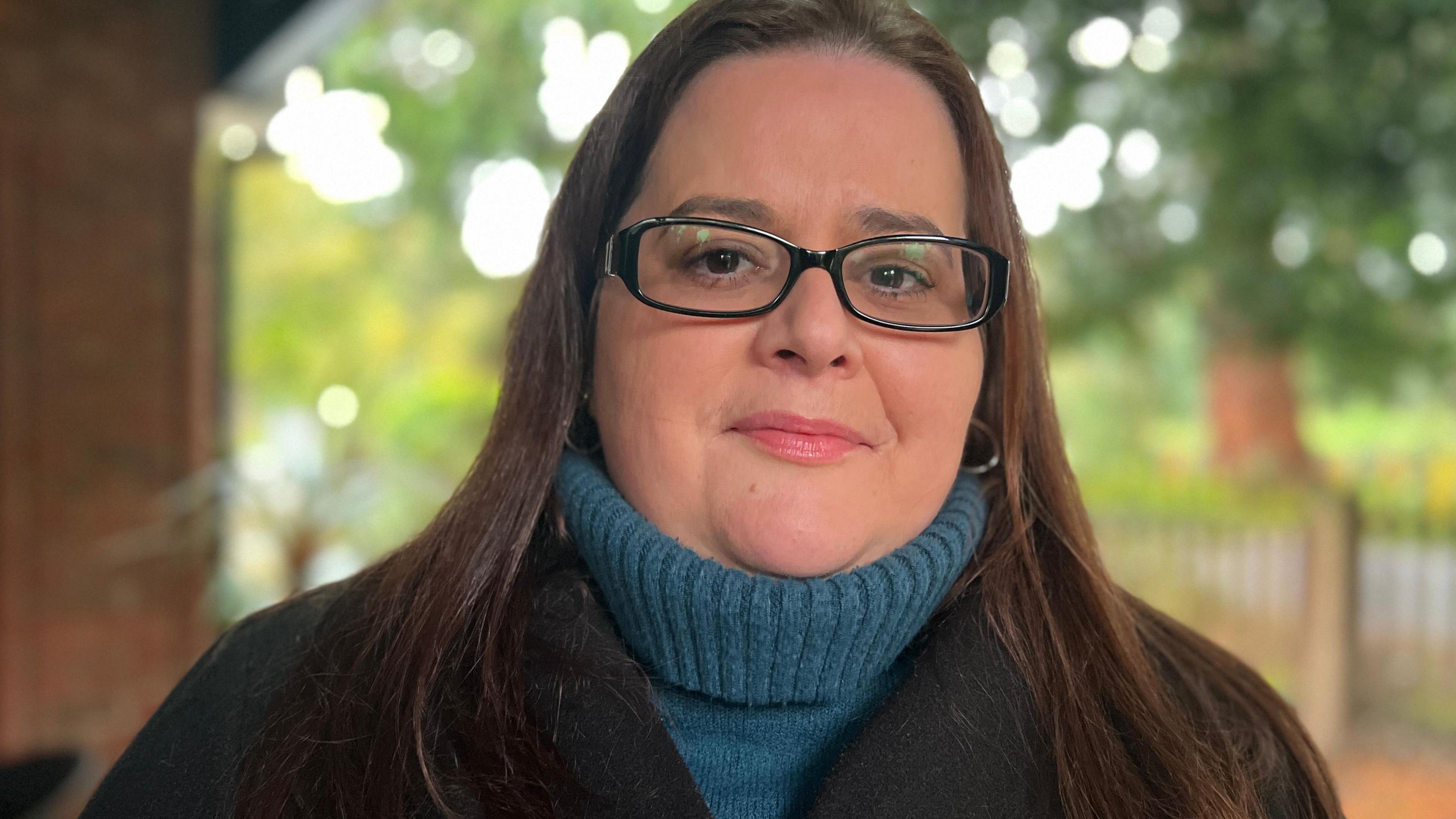 Katy Cole, who has long mid-brown hair falling over her shoulders and is wearing black-framed glasses. She is wearing a mid-blue roll-neck chunky jumper under a black coat. She is standing outside and behind her are blurred out trees, grass and a fence.