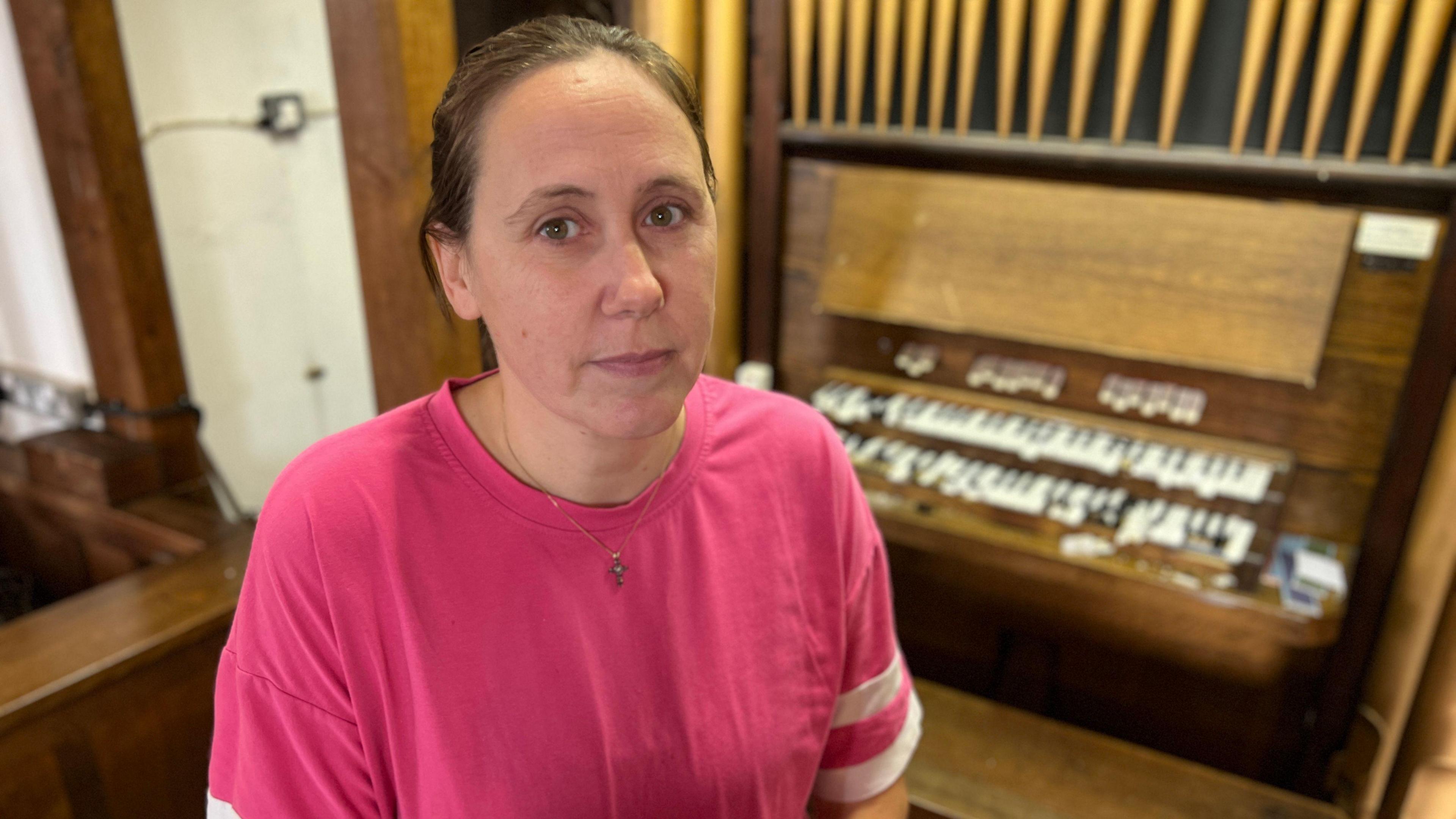 Woman with hair tied back wears a pink t-shirt. She stands in front of a broken and smashed church organ. She looks sad.