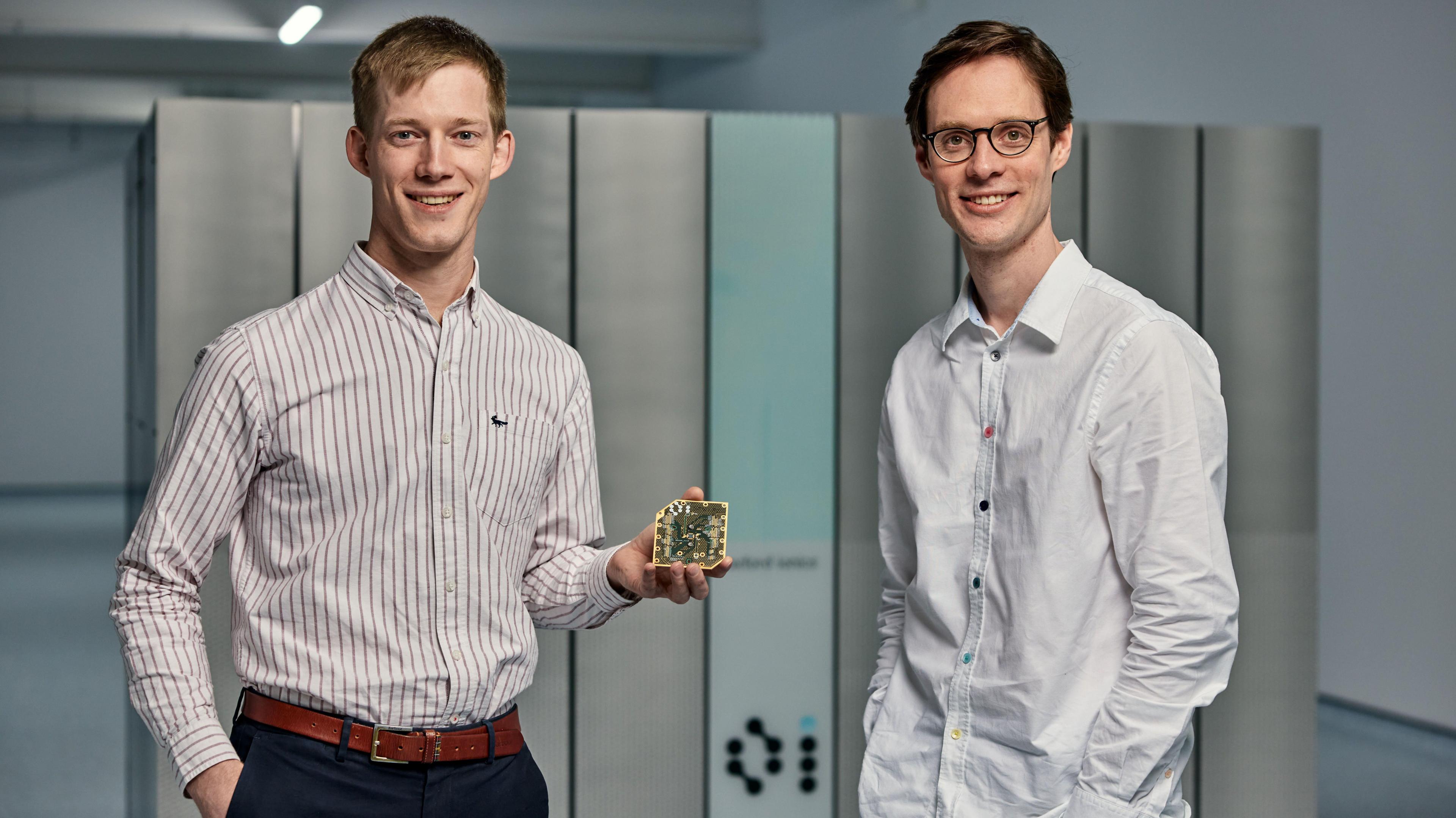 Dr Chris Ballance and the other Oxford Ionics co-founder, Dr Tom Harty, are smiling for the camera in front of Quartet. Dr Ballance is holding a chip.