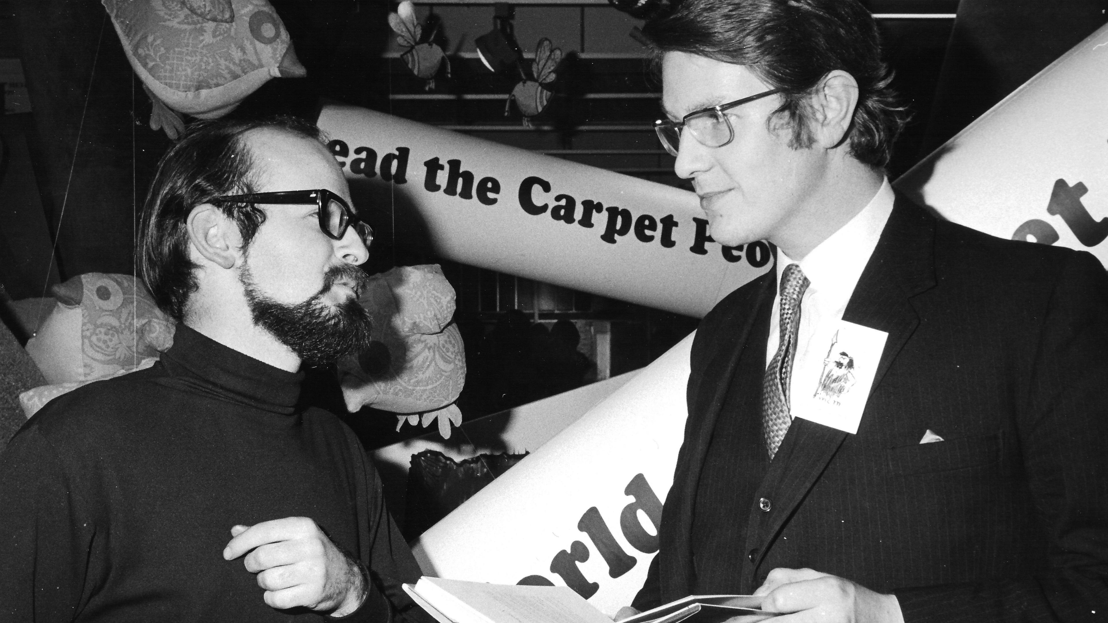 A black and white photo of two men facing each other. On the left is a shorter man with a dark hair and a dark beard wearing glasses. On the righ tis a taller man with thick hair and dressed in a suit. A banner behind them says "read the Carpet People"