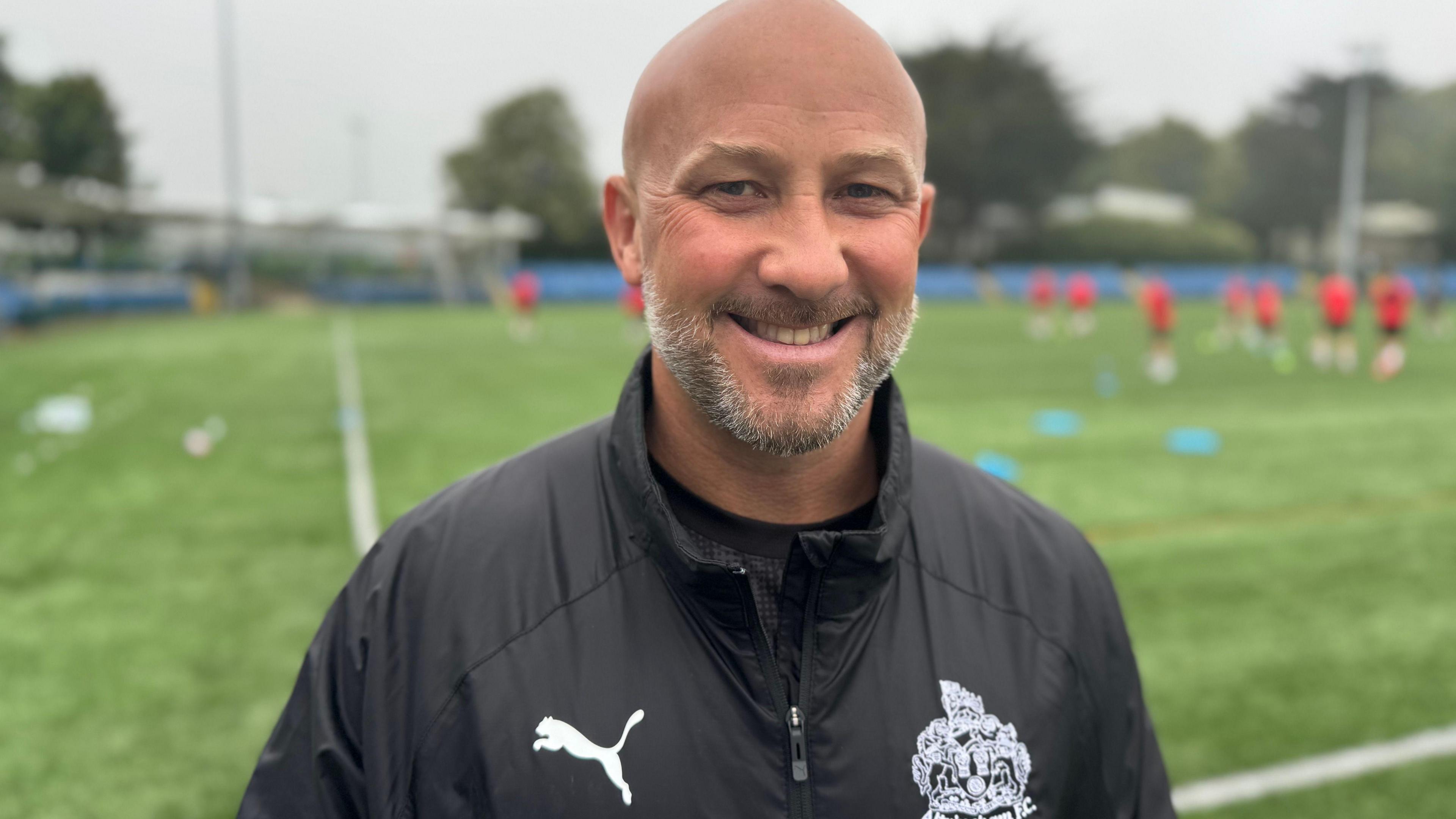 Phil Parkinson is bald with a grey beard, he smiles and wears a dark rain coat. He stands by a green football pitch with players wearing red blurred out behind him.