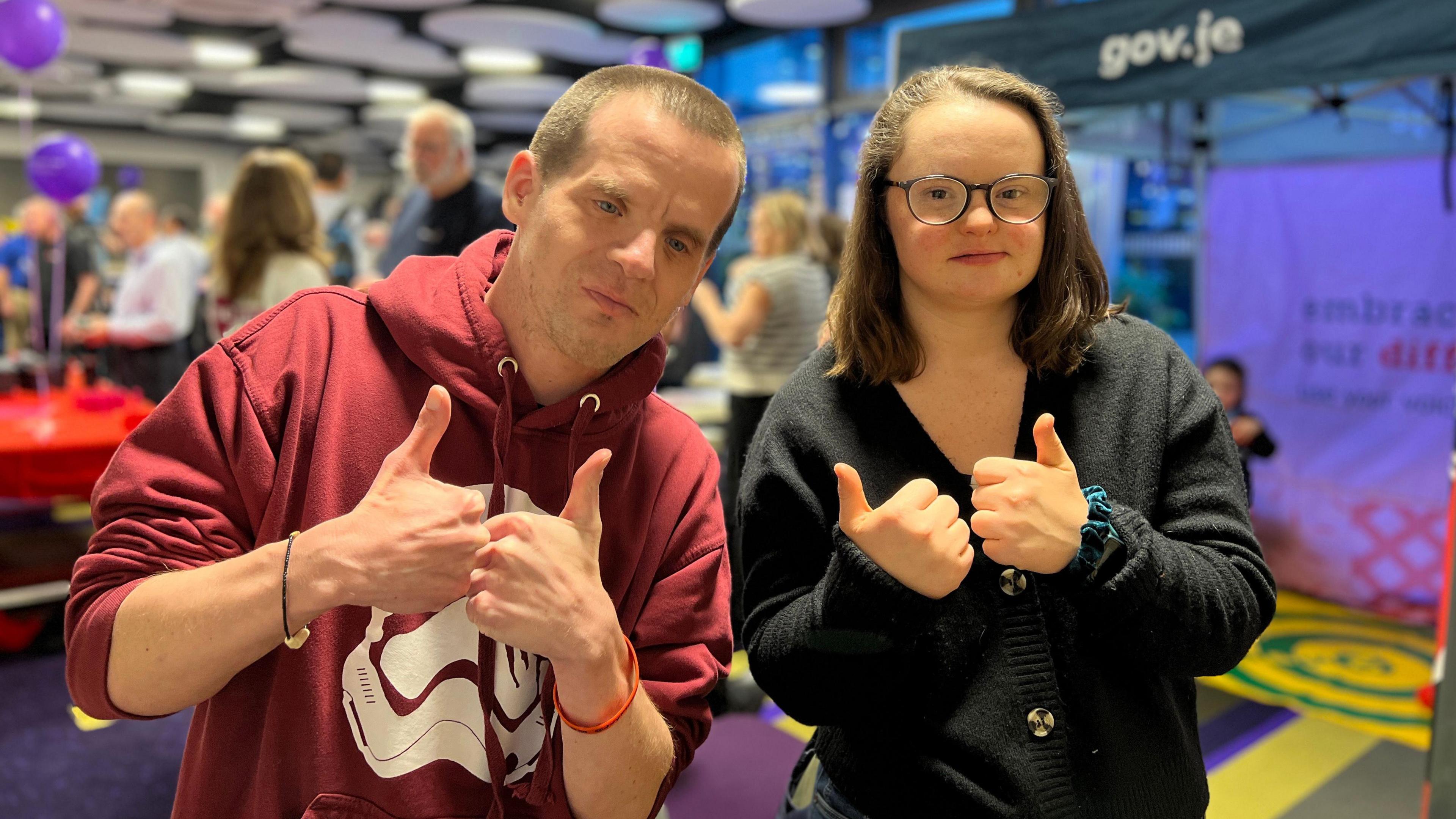 Matt and Rachel look at the camera and give a thumbs up with both hands. Matt is wearing a dark red hoodie with a stormtrooper mask on the chest and Rachel has a dark blue jumper on. Matt has shaved hair and Rachel has dark hair down to her shoulders. Behind them people are standing around stalls.