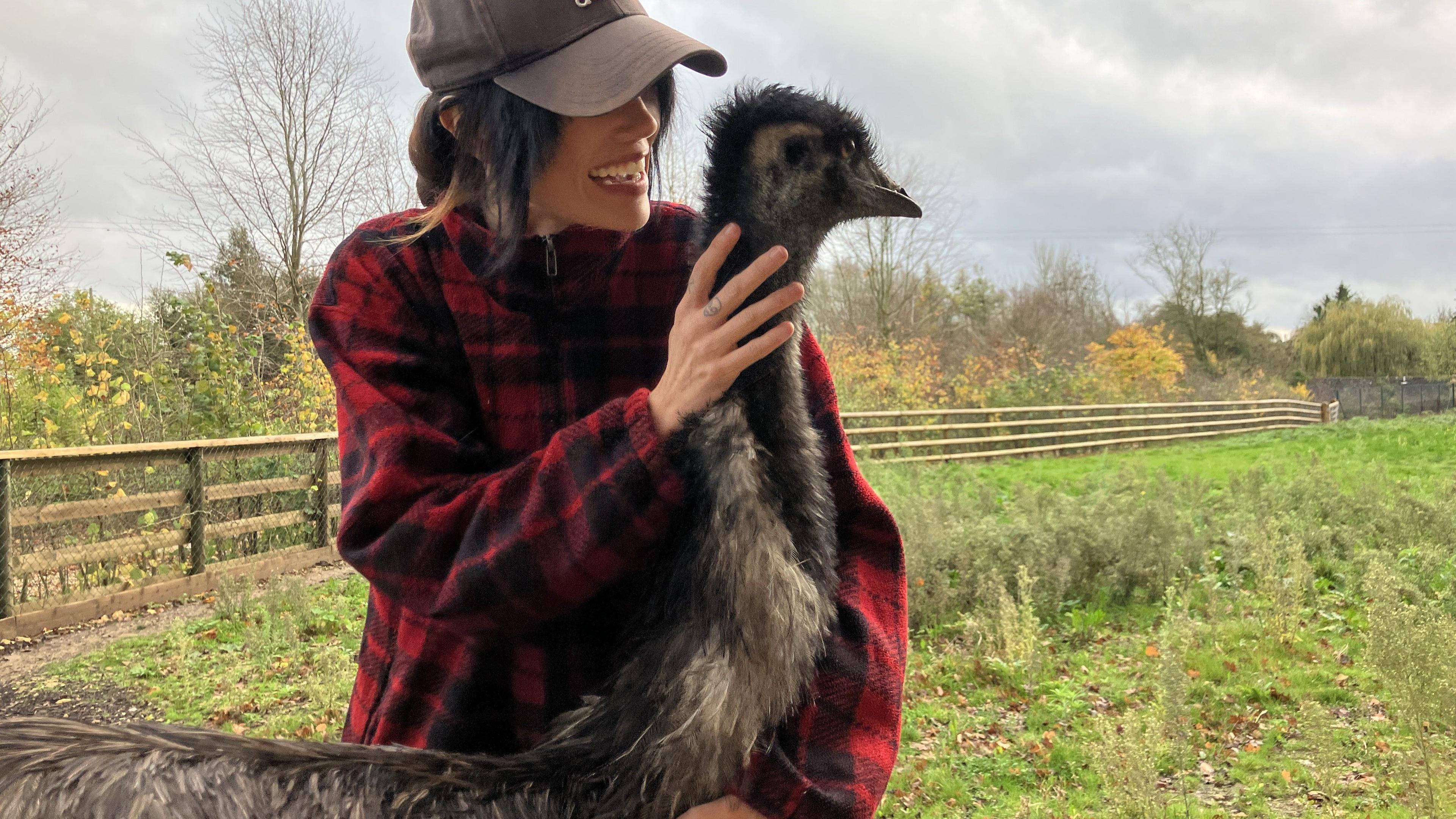 Rhi Evans cuddles an emu in a field. Ms Evans is wearing a black baseball cap and a red plaid shirt.