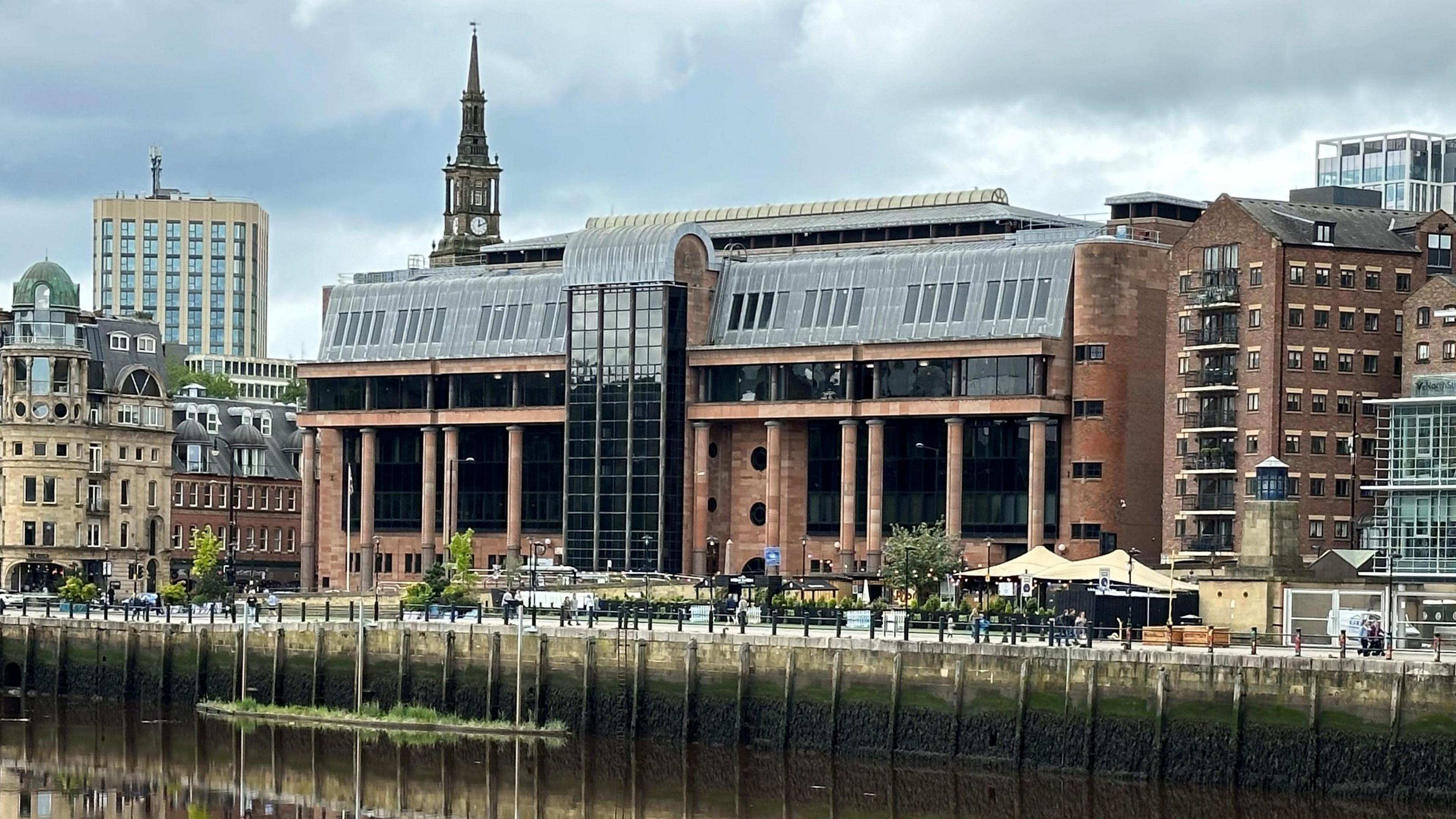 Newcastle Crown Court reflected in the River Tyne running in front of it. It is an imposing building made from smooth red stone with massive black windows and tall columns along its frontage.