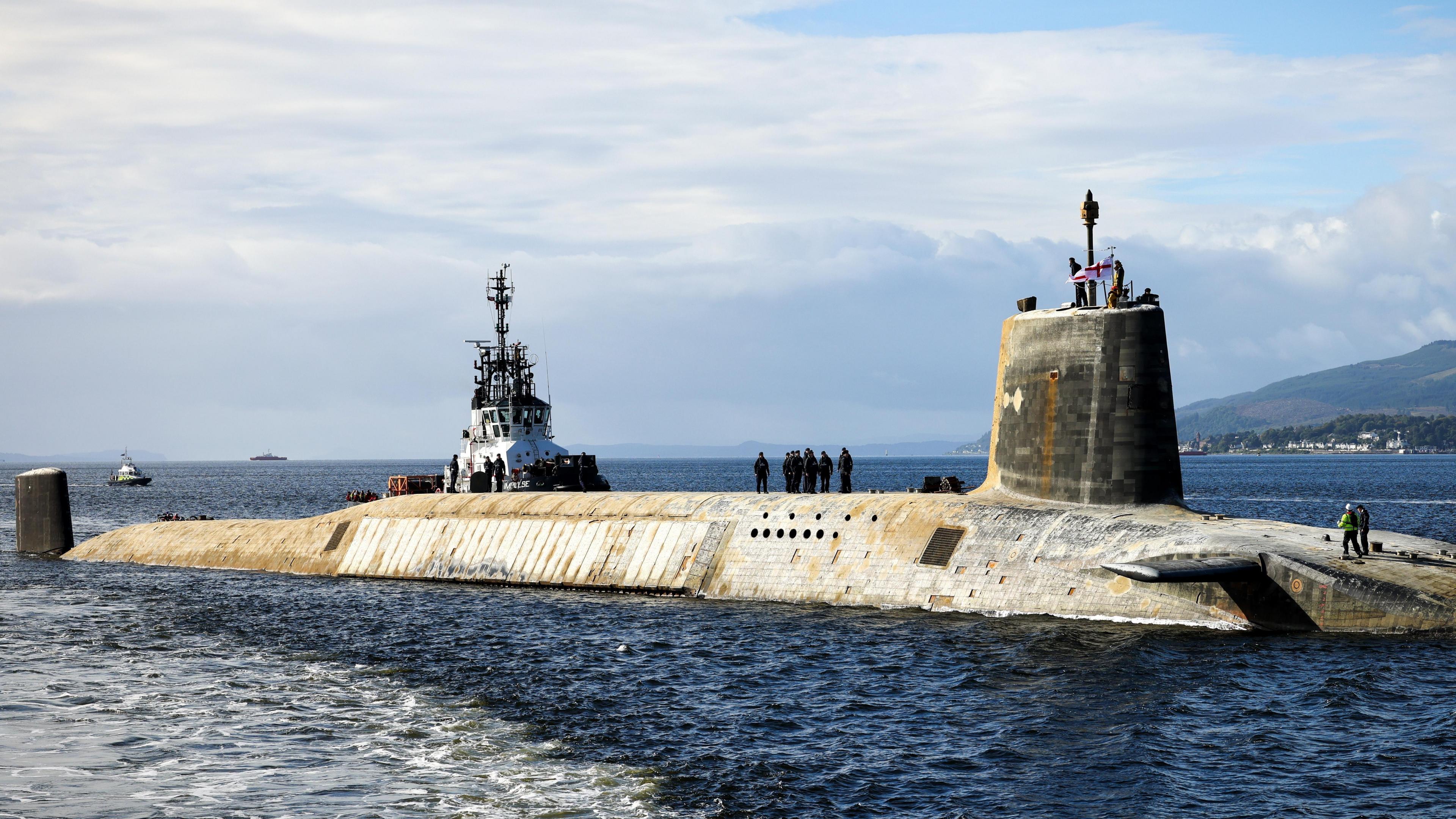 A British Navy Vanguard submarine seen on the surface from a distance, with a number of personnel on deck, some holding flags and others in uniform