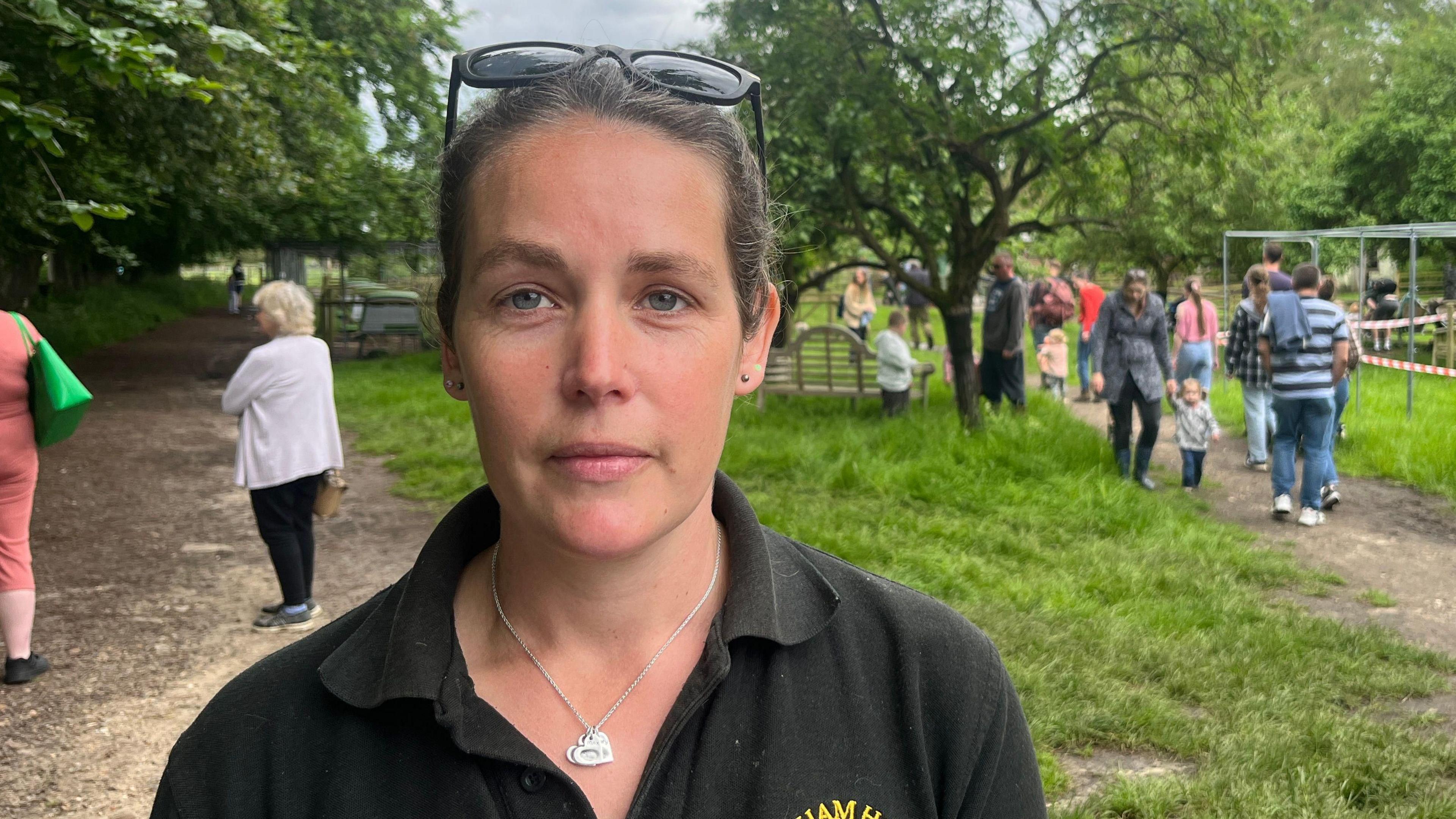 Kate Ockelton smiles at the camera. She has dark hair that is tied back behind her head and she wears a dark green polo top with sunglasses on her head and a necklace around her neck.