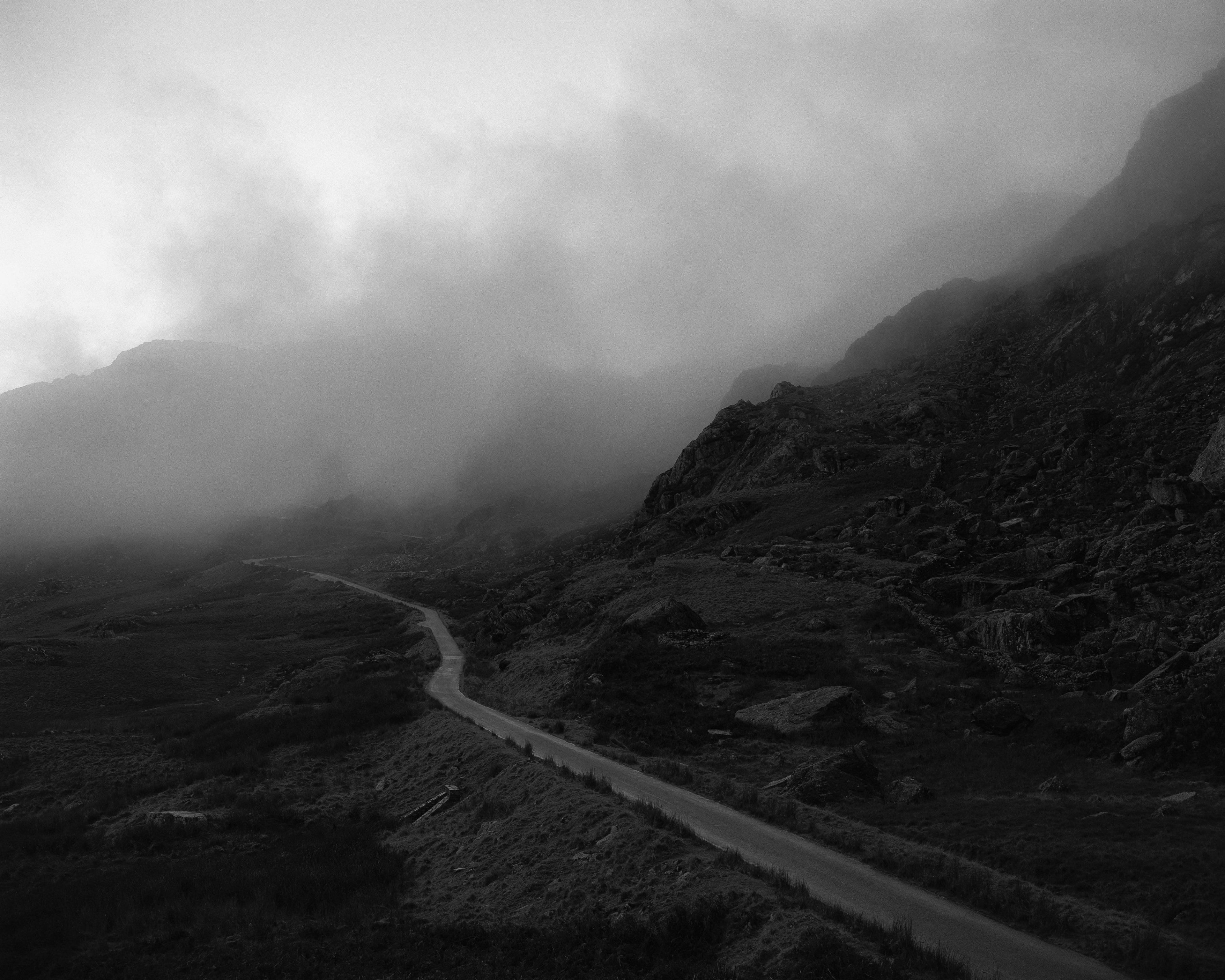 Kyle's black and white photograph of a mountain road with a craggy hillside on one side and grass sloping bank on the other. The sky above is misty white. 