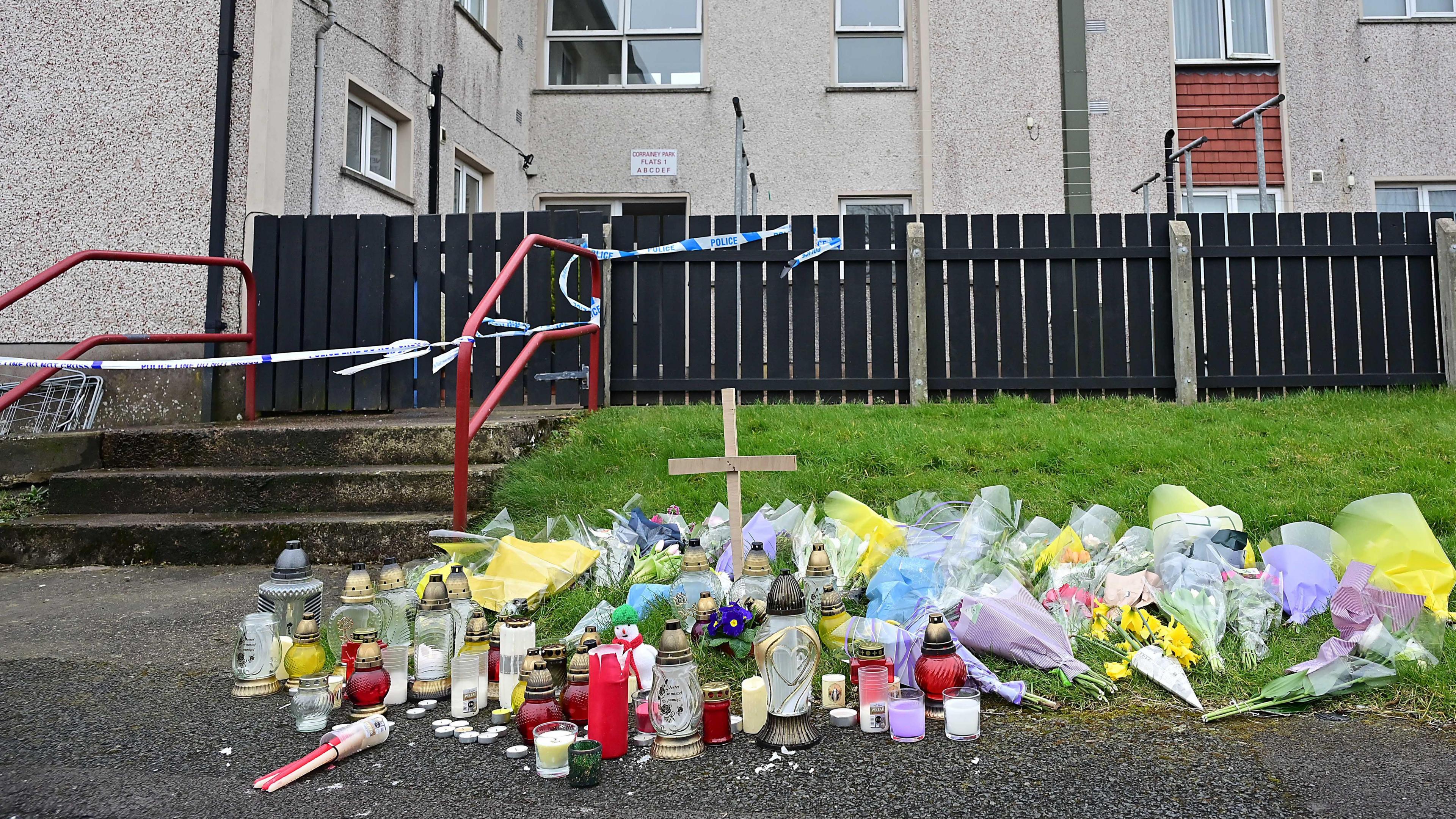 Candles, flowers, and a cross left on the ground outside a block of flats with the entrance covered by police tape