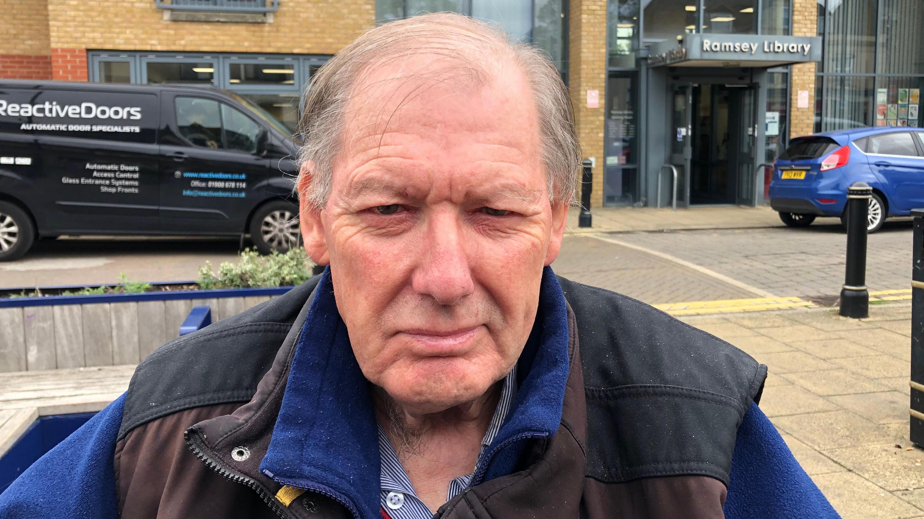 Roger Papworth is 76, has thinning combed over wispy dark and in places grey hair. He is wearing a stripped shirt, with a blue fleece and a brown body warmer and he is sitting in front of Ramsey town library with vehicles parked outside