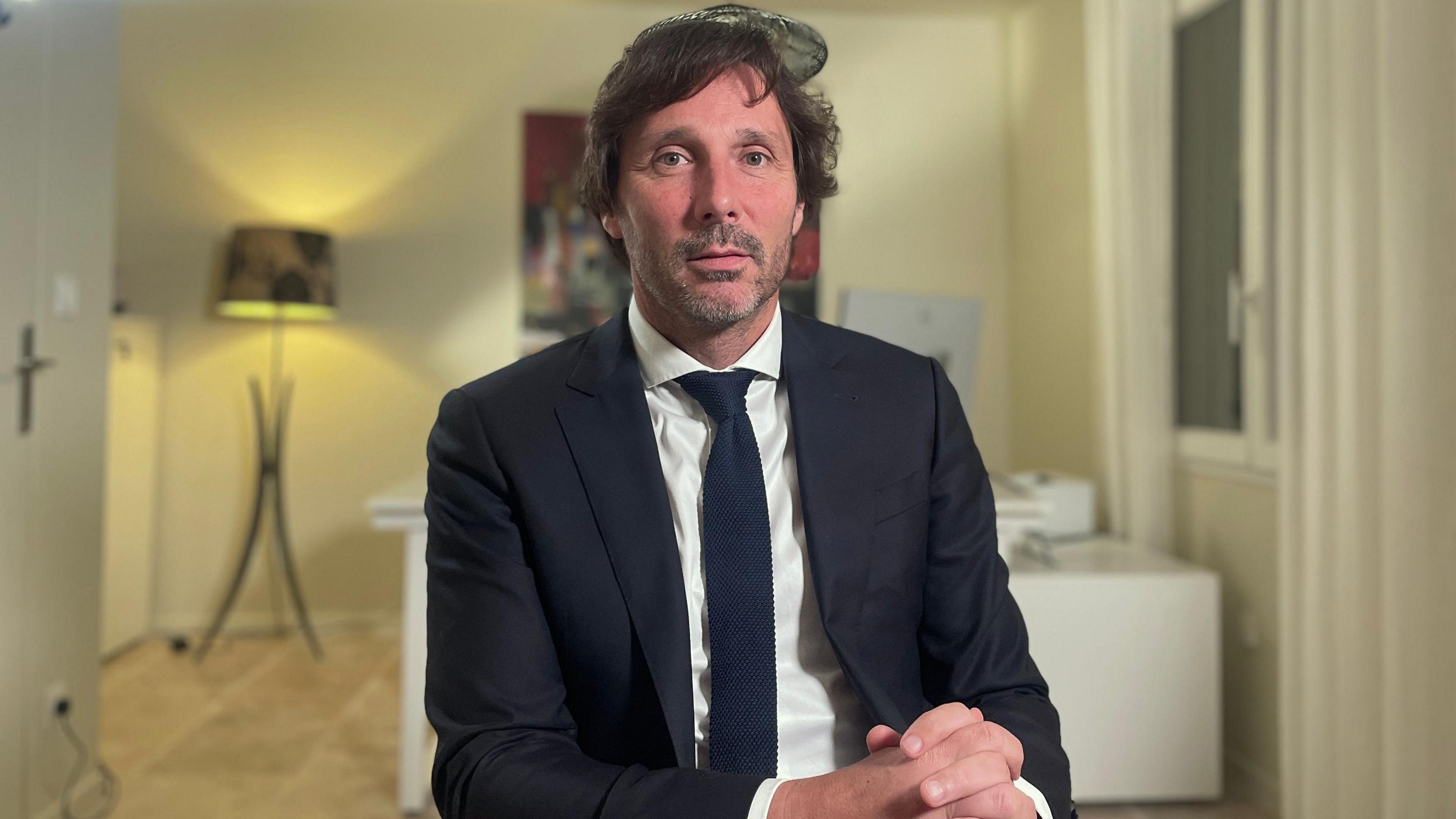 Dr Laurent Layet, a man with dark brown hair and stubble, wearing a dark suit and a dark tie on a white shirt, sitting with his hands clasped on his knee in a liveing room, with art on the walls and a lamp in the background.