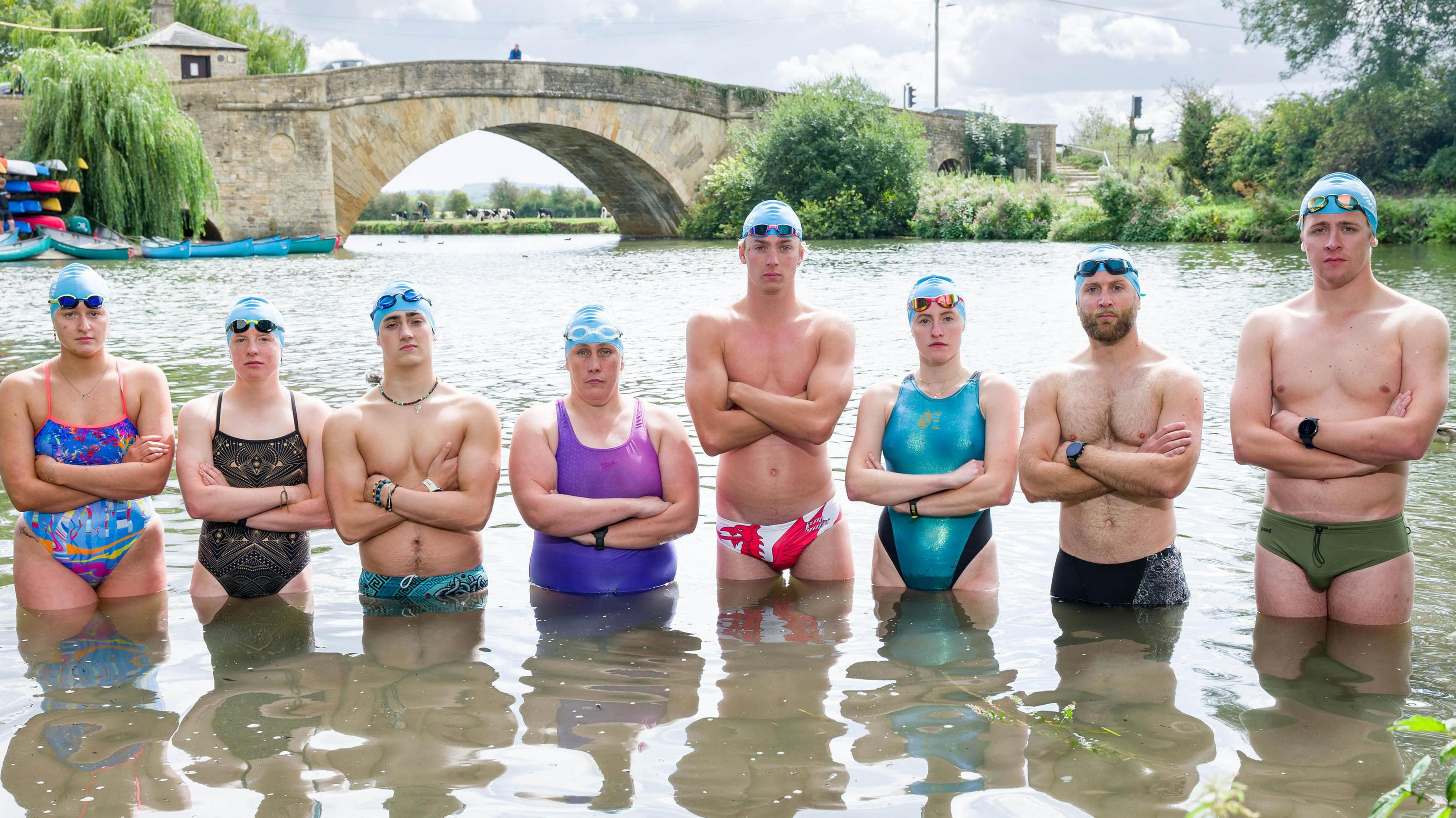 Eight swimmers stand waist deep in water, four male, four female, ready to begin the challenge. They have swimming caps and goggles on their heads, their arms are crossed. A stone bridge crosses the river behind them.