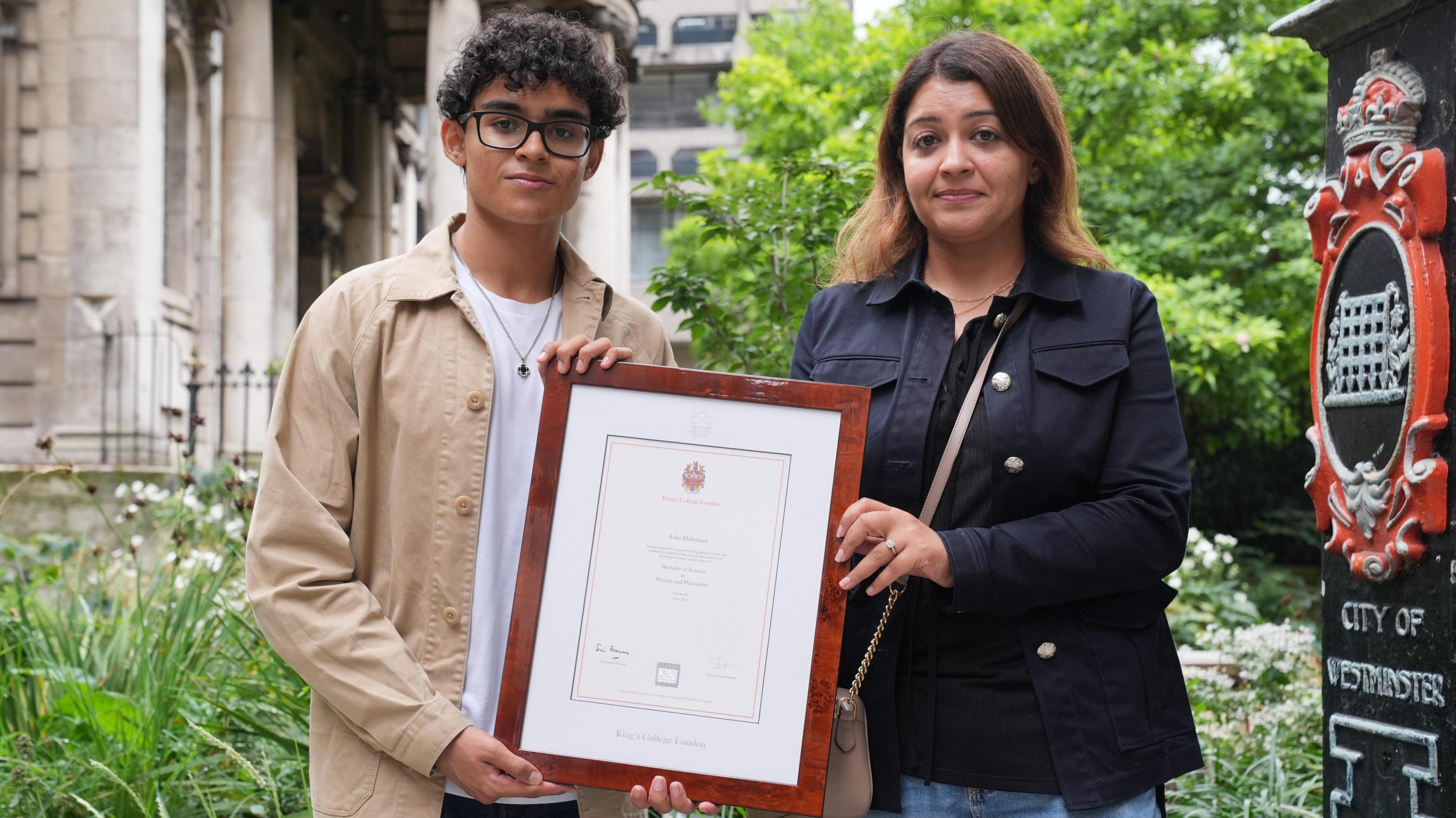 Zain is wearing a white top and beige jacket. He has short curly hair and is wearing glasses. He is stood beside his mother, who is in a bloack top and navy jacket. They are holding a university certificate and looking towards the camera.