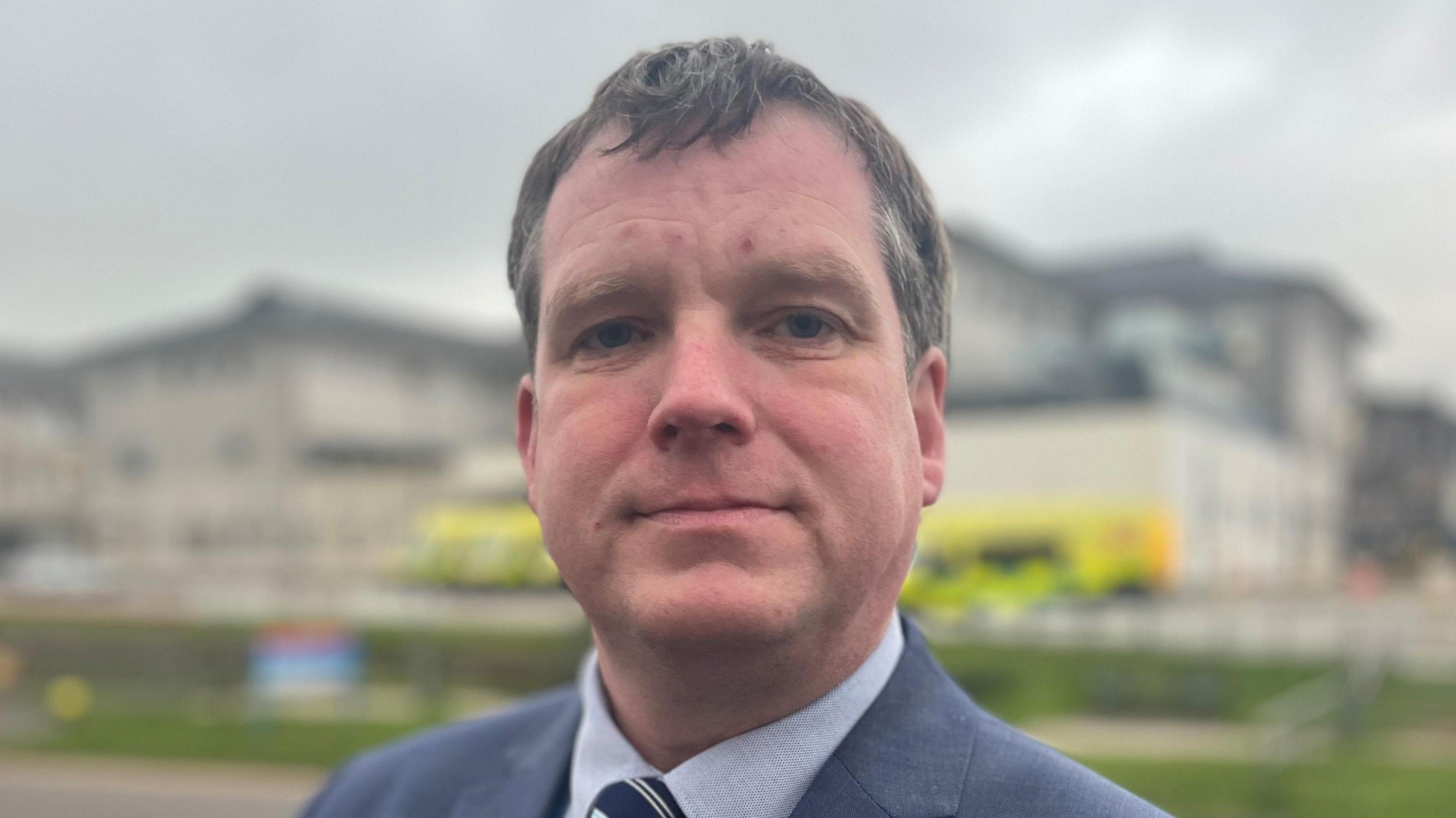 Head and shoulders of a man with grey hair wearing a grey shirt and black striped tie, with a blurred image of the Royal Cornwall Hospital and ambulances behind 