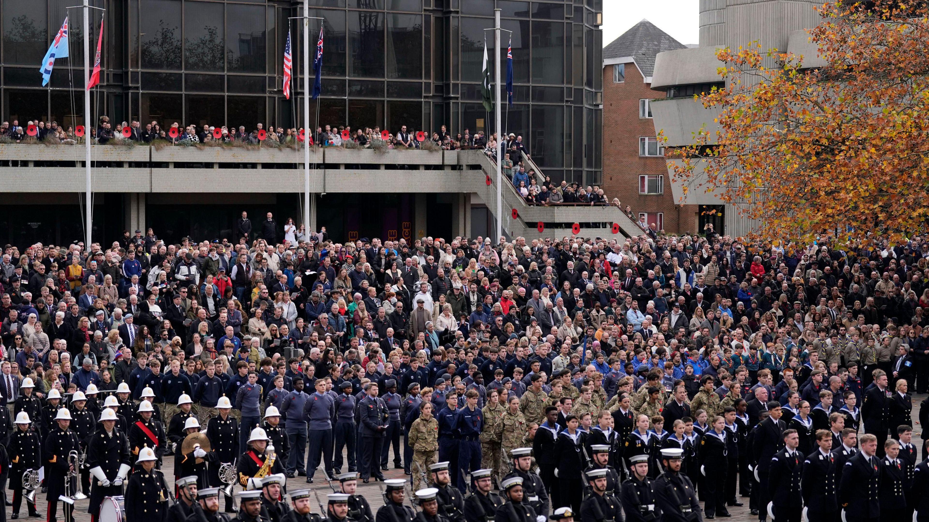 A large crowd in Guildhall Square. Military personnel stand to attention in orderly rows, surrounded by spectators on ascending steps and upper level walkways