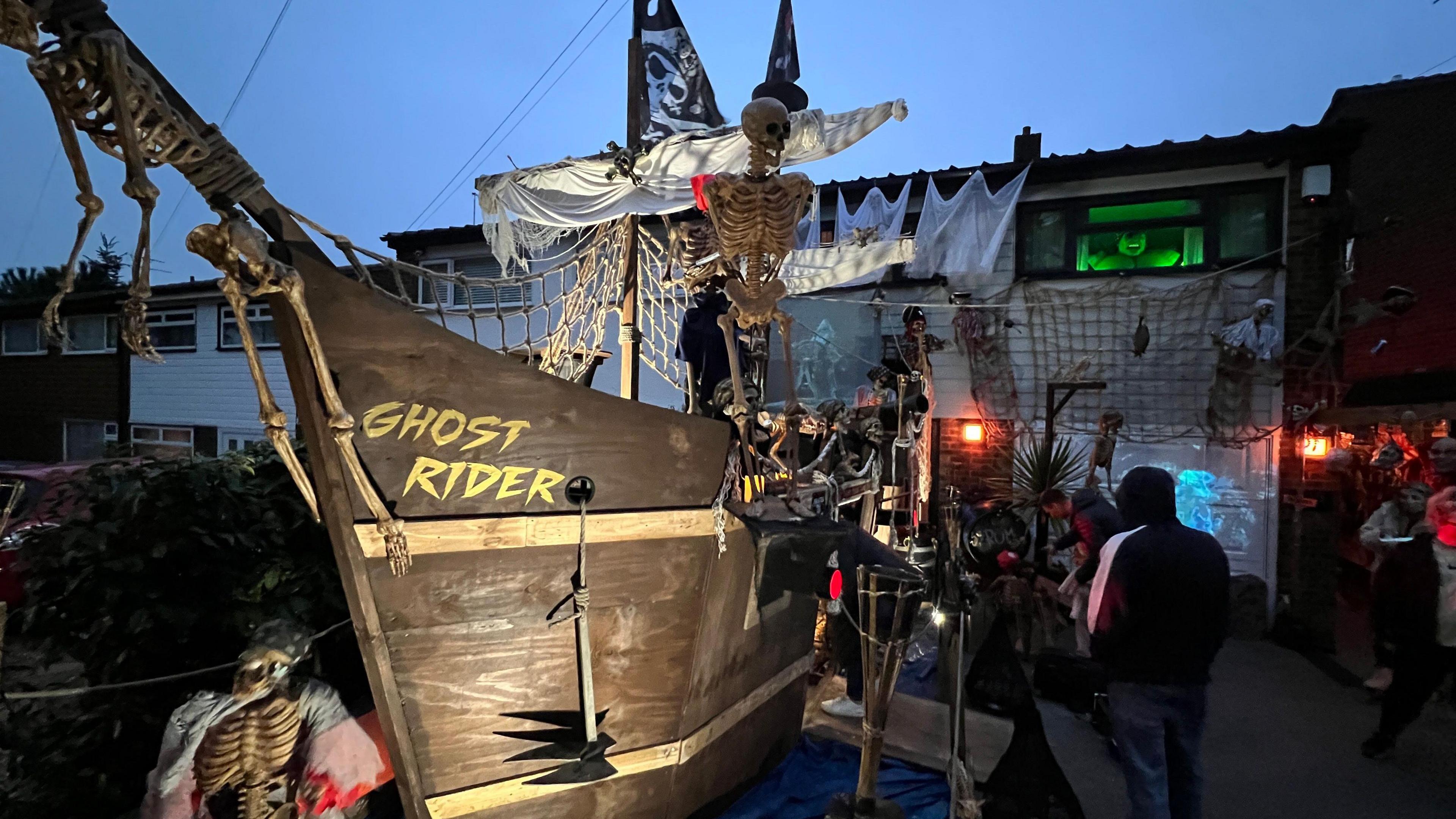 A pirate ship decorated for Halloween. It is brown with "Ghost Rider" written across the front in yellow text. There are three skeletons hanging from the ship, which is dimly lit at dusk.