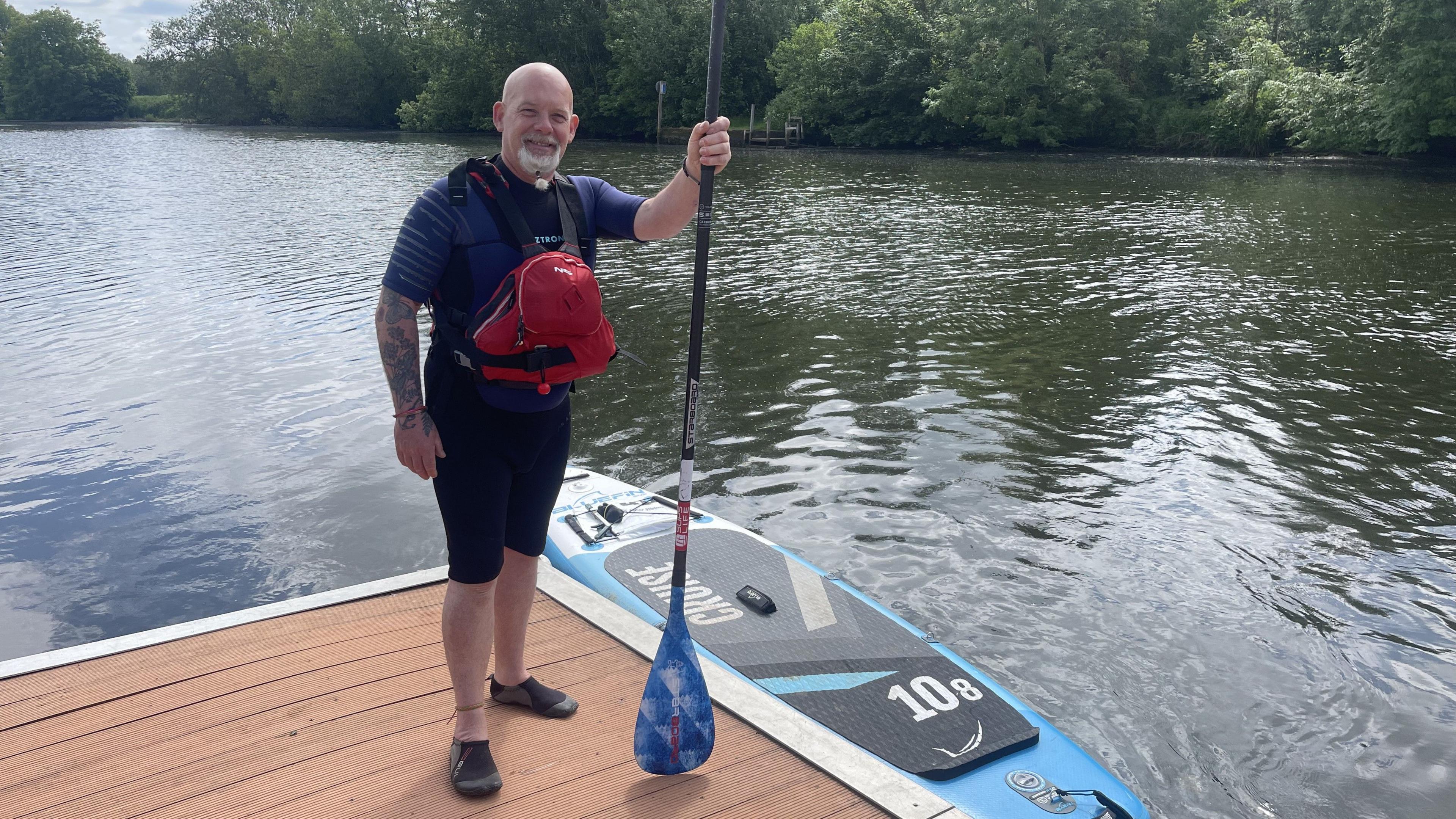 Darren Reeve - a bald man with a goatee beard - stands on a jetty with a paddle in his hand. There is a paddleboard in the river next to him. The wide river is lined by low-hanging trees.