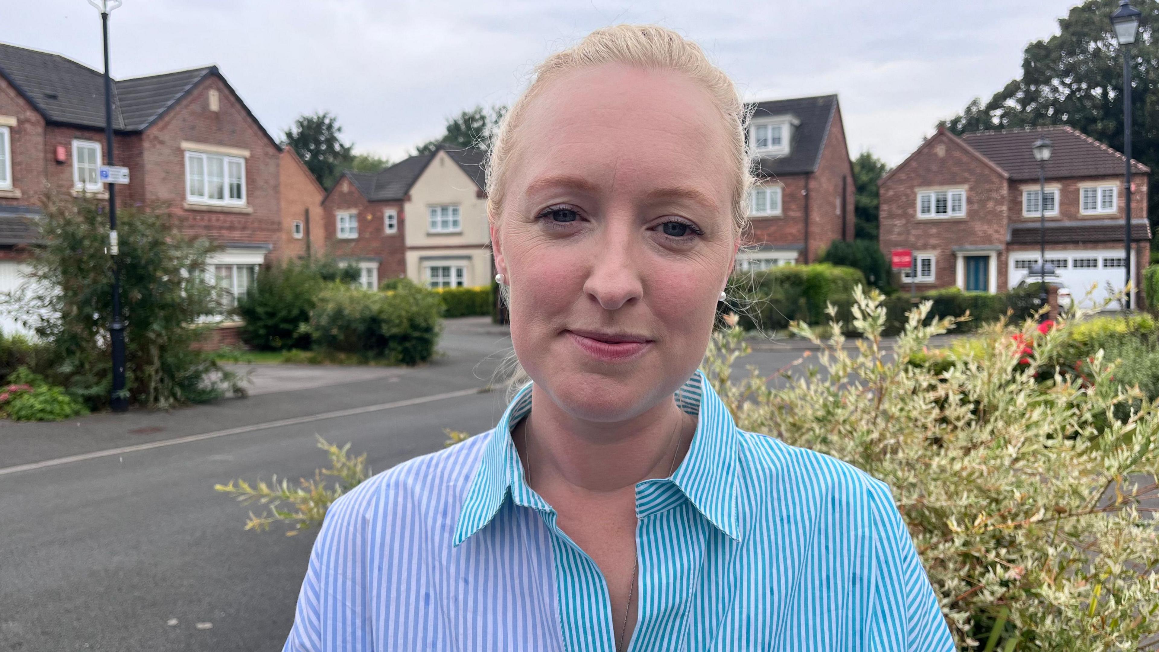 A woman with blonde hair, pulled back into a ponytail, wears a blue striped shirt and pearl earrings. She is stood outside houses on a quiet residential street.