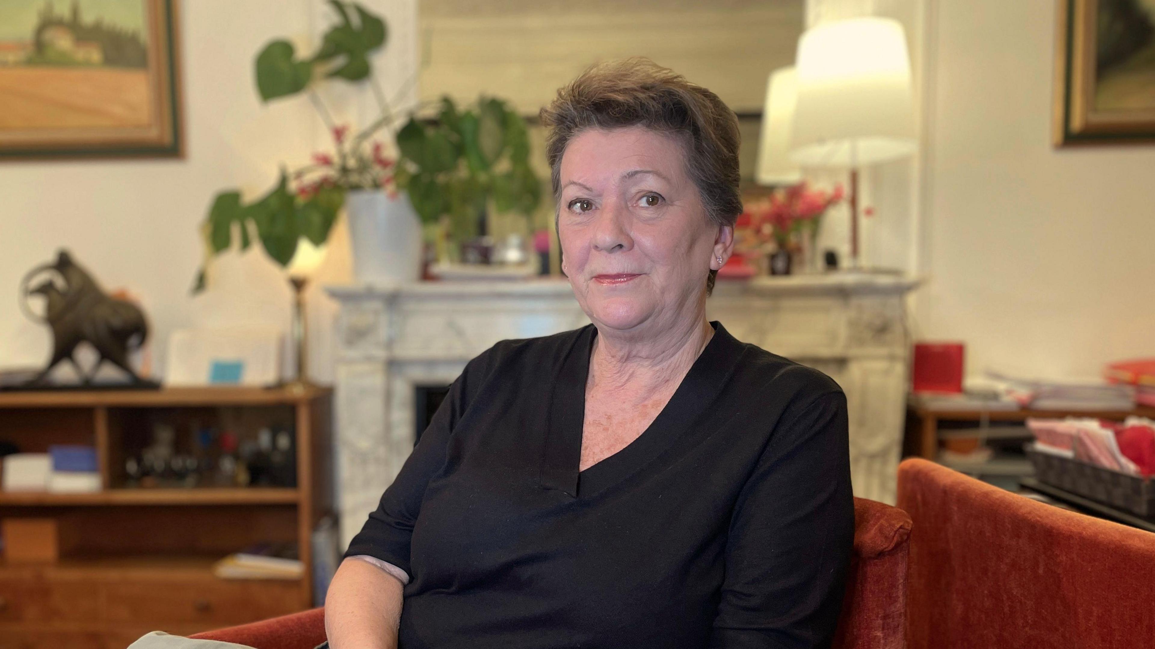 Florence Rault, a woman with short brown hair, wearing a black top and looking straight at the camera as she shits in an armchair in a sitting room, with paintings on the wall and a sculpture on a sideboard, with a large pot plant and soft lamps in the background.