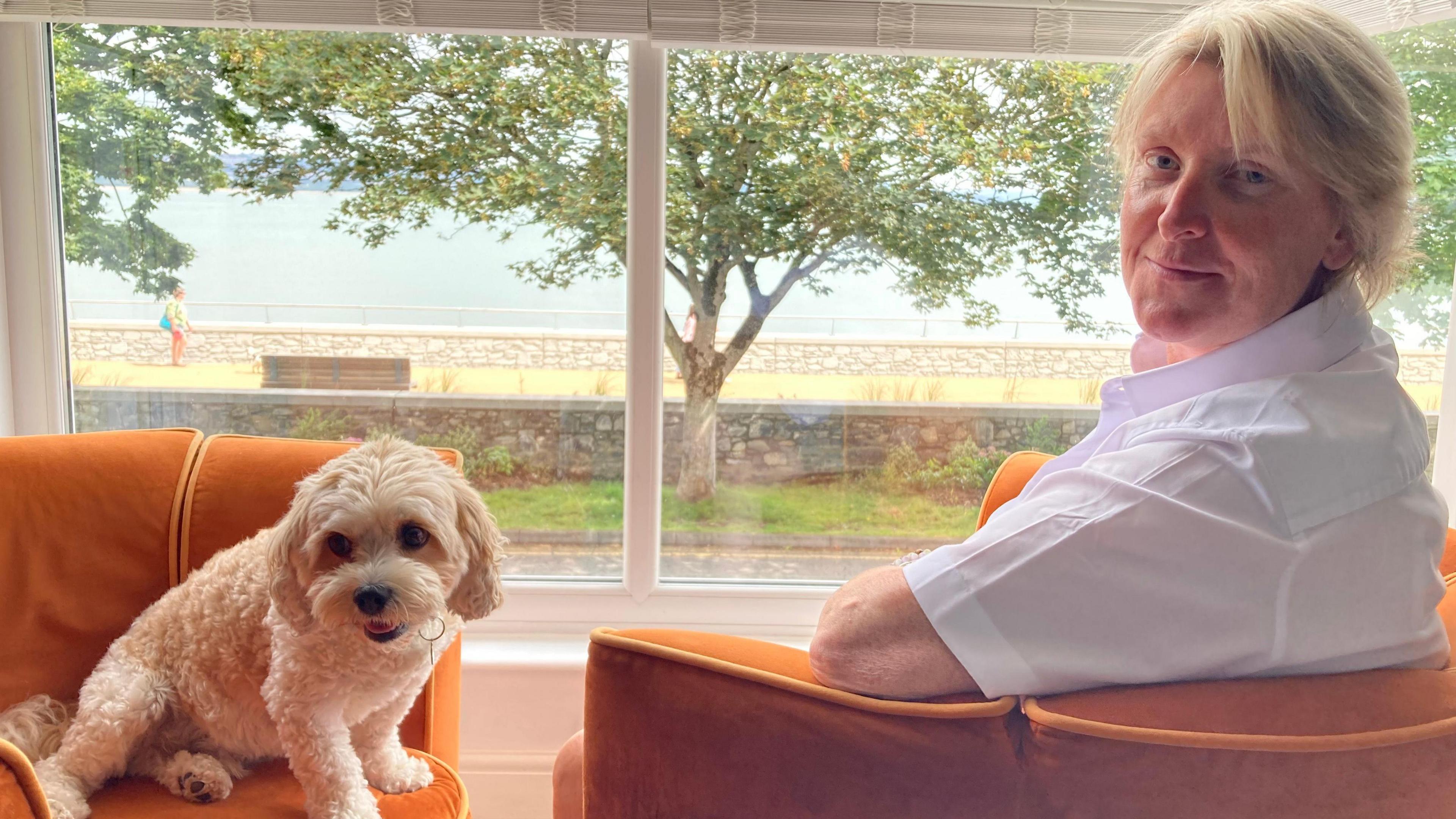 A woman with short blonde hair, wearing a white shirt, is smiling at the camera. Next to her is a small white dog. They are both sat on orange chairs, and behind them is the promenade. 