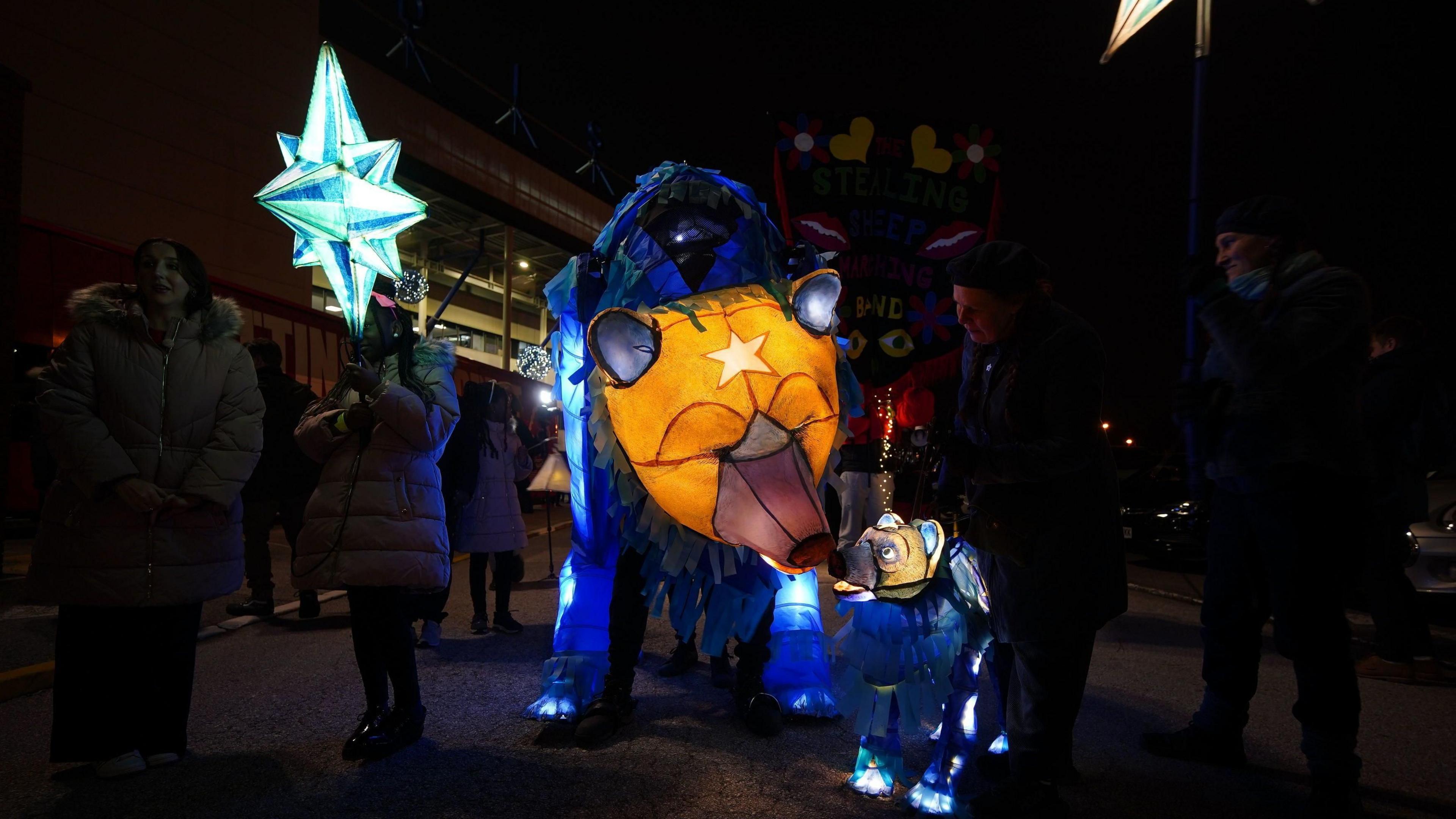 A lit-up lantern bear next to a smaller one, with people holding lit up stars in the dark