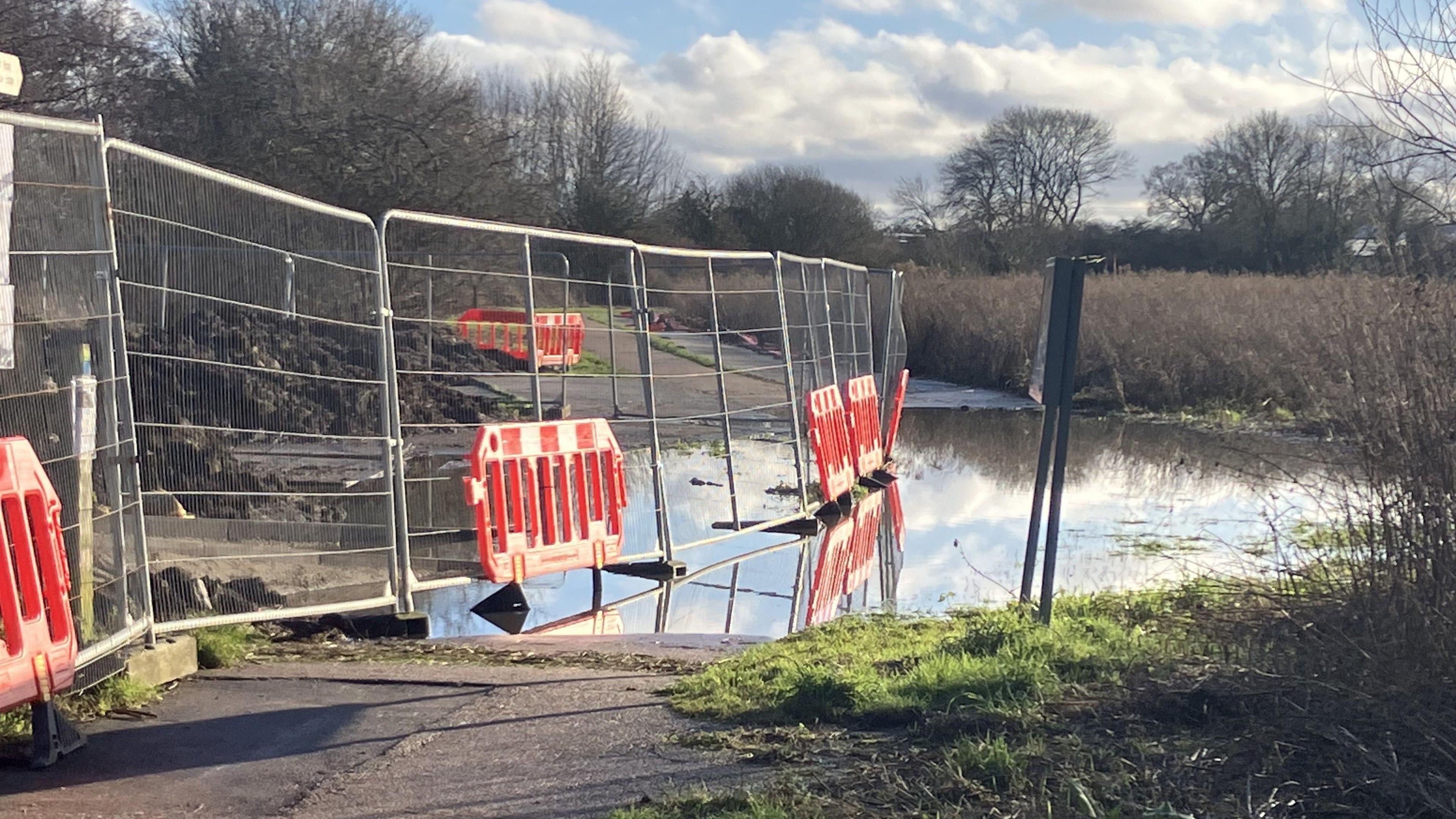 A high metal fence with red bollards attached borders a flooded pathway close to the bridge in a rural setting 