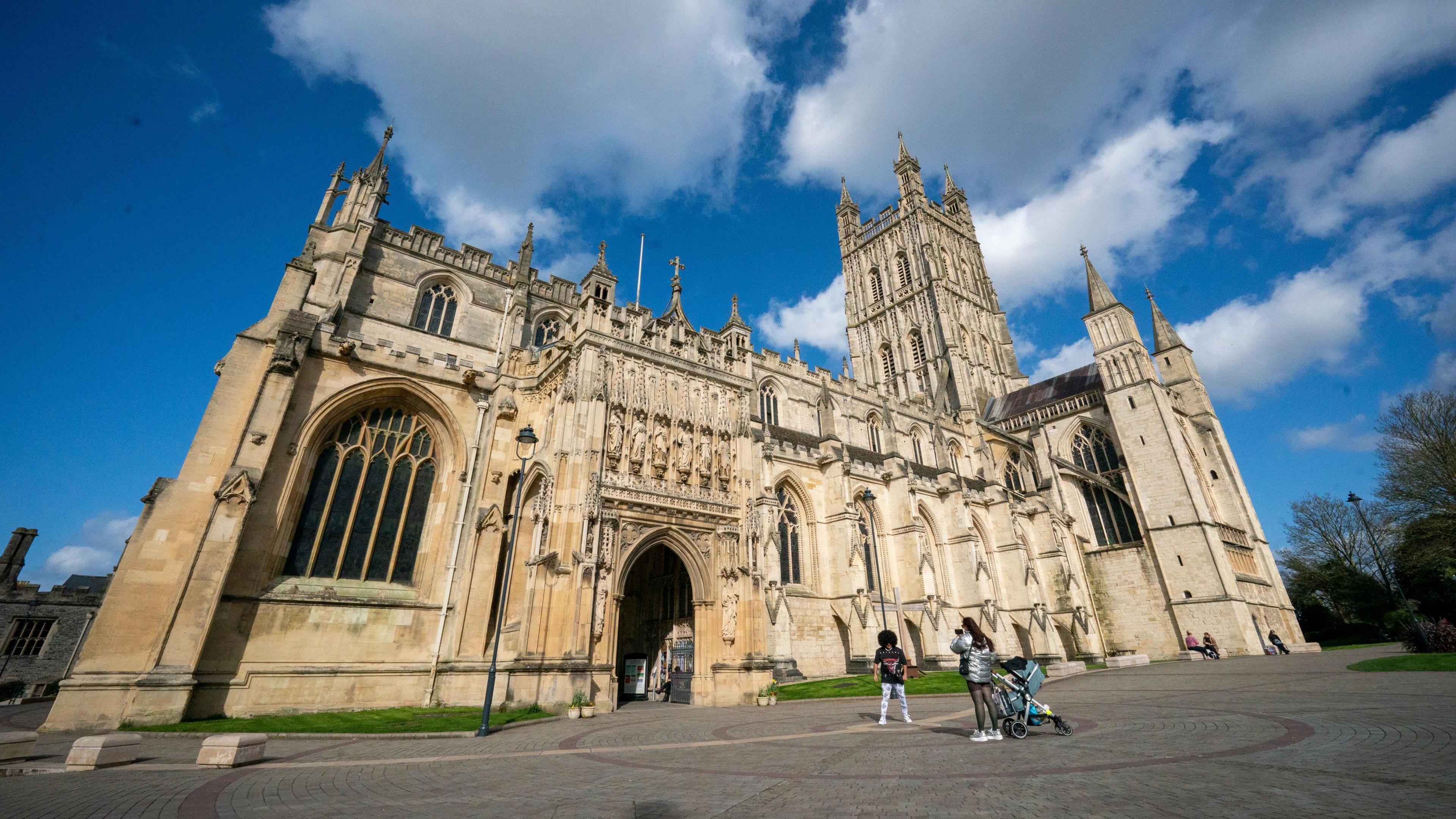 A cathedral pictured with people standing outside posing for photos under a blue, cloudy sky