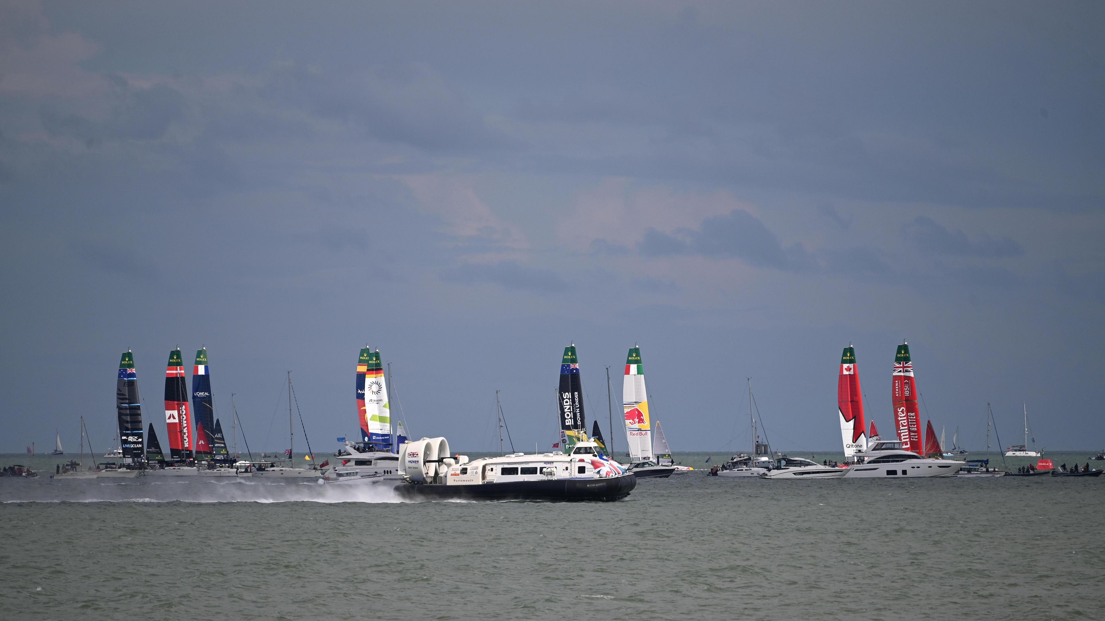 Dark skies above grey seas surround several high speed racing yachts involved in a competition. A passenger hovercraft passes through the foreground.
