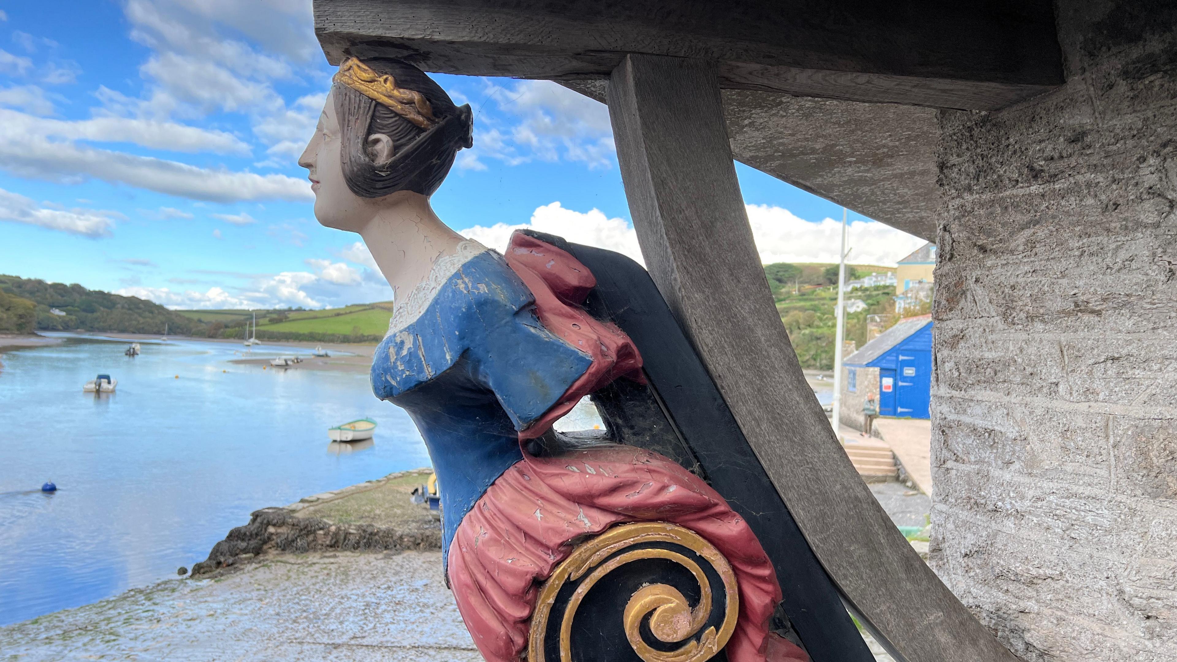 Ship figurehead of a young woman with a blue top and red skirt and her brown hair tied up, with a river in the background and a curved wooden post emerging from a building behind.